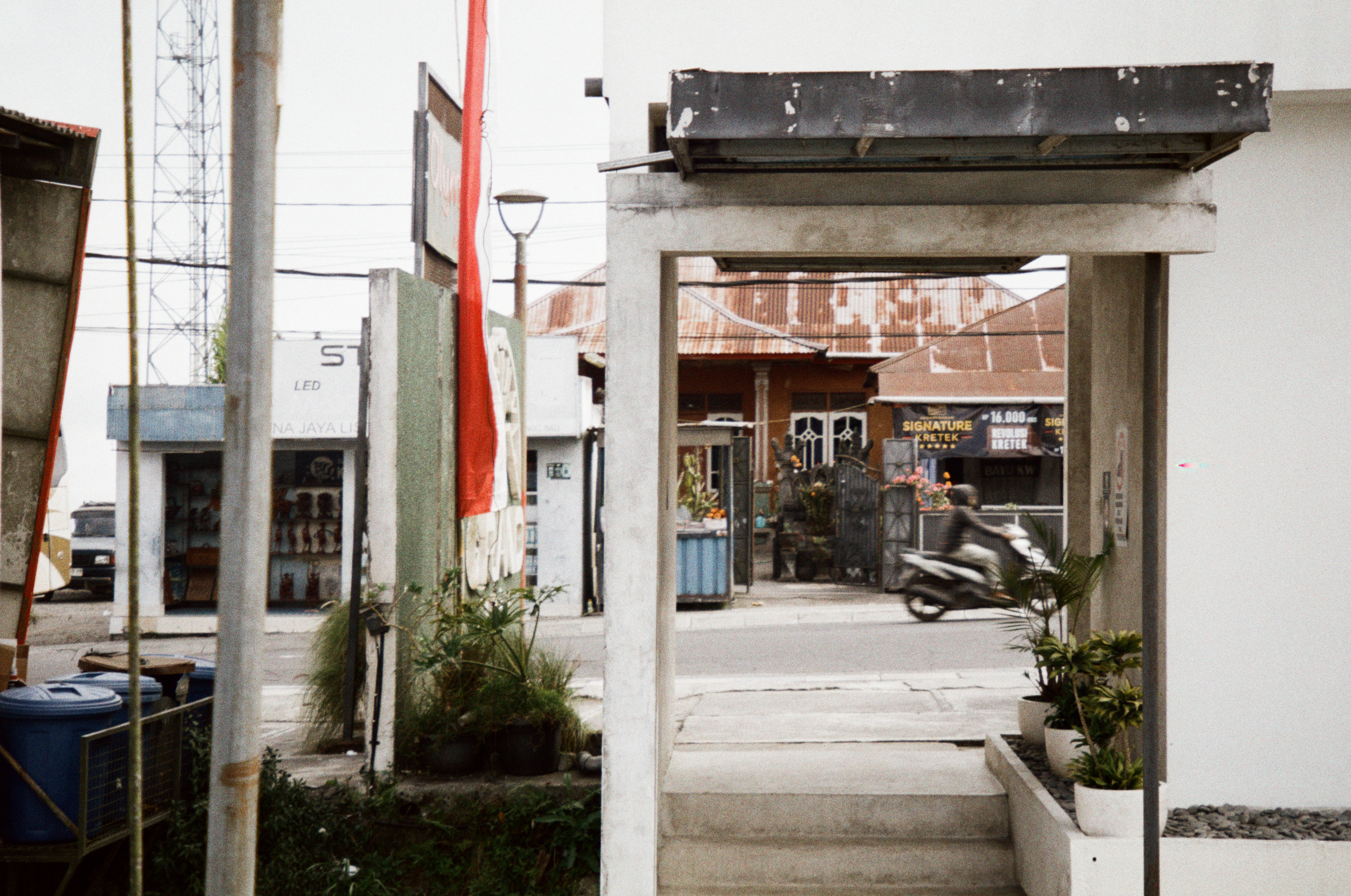 A rider on a scooter passes beyond a concrete gateway, with shopfronts, power lines, a lamppost, and a red-and-white banner along the street.