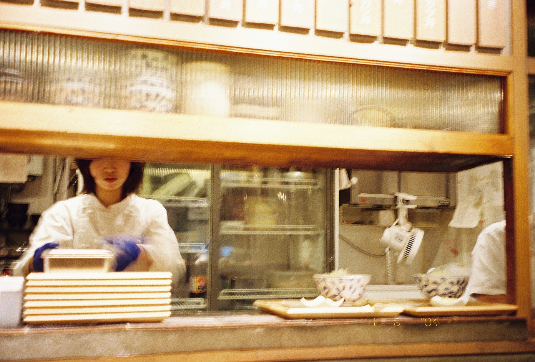 A cook in white with blue gloves reaches across a kitchen counter stacked with lacquered trays and patterned bowls behind a service window.