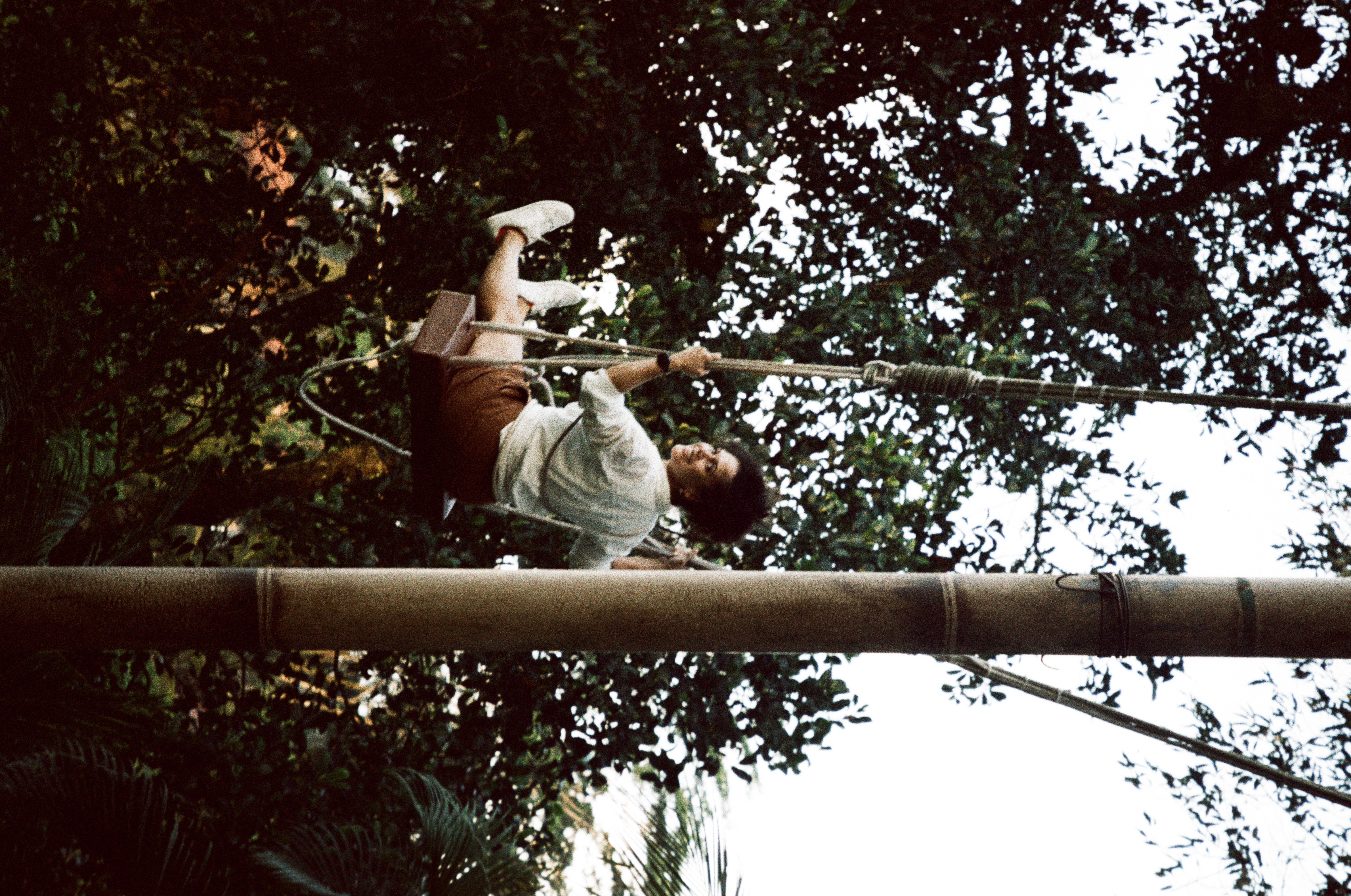 A smiling person in a pale shirt and brown shorts sits on a rope swing beside a tall pole amid dense foliage.