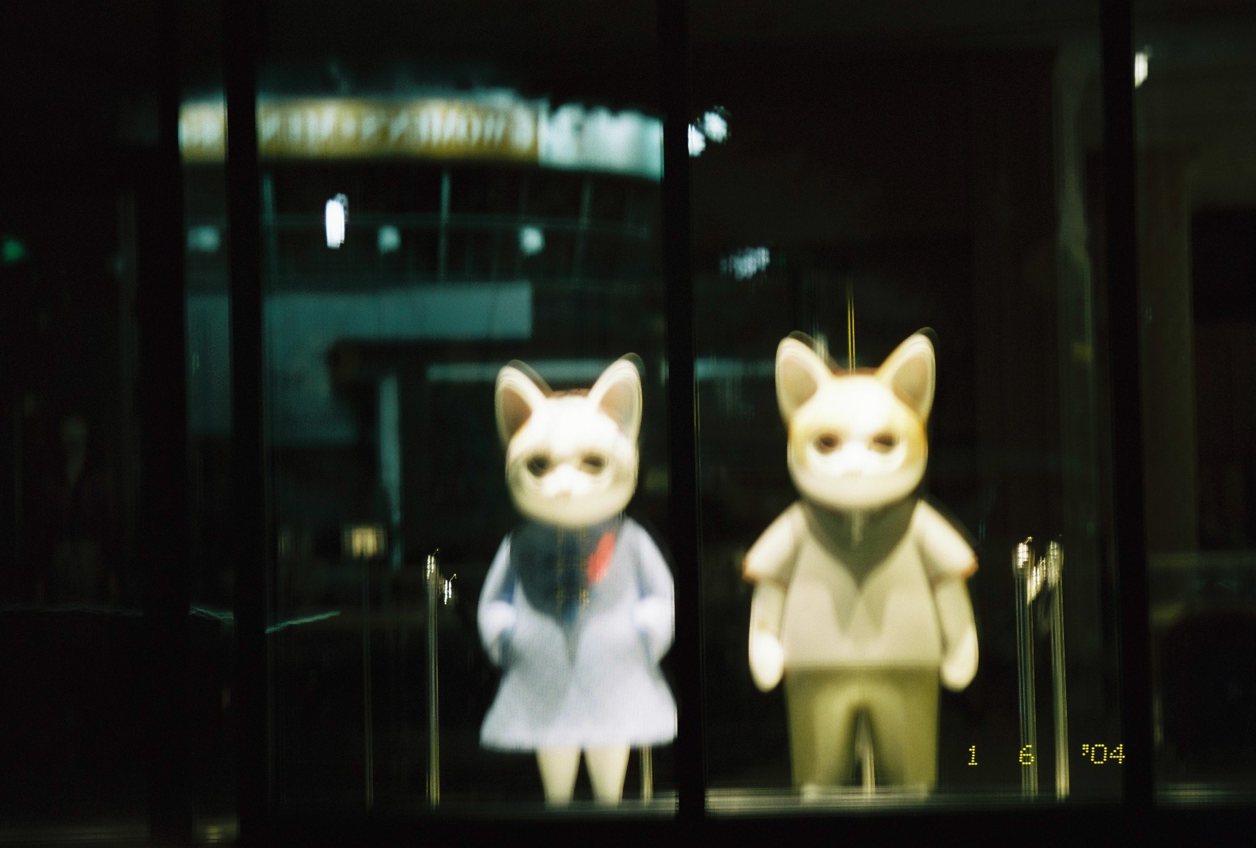 Two pale, cat-eared mascot figures stand behind a dark shop window at night, one in a light dress with a red collar accent.