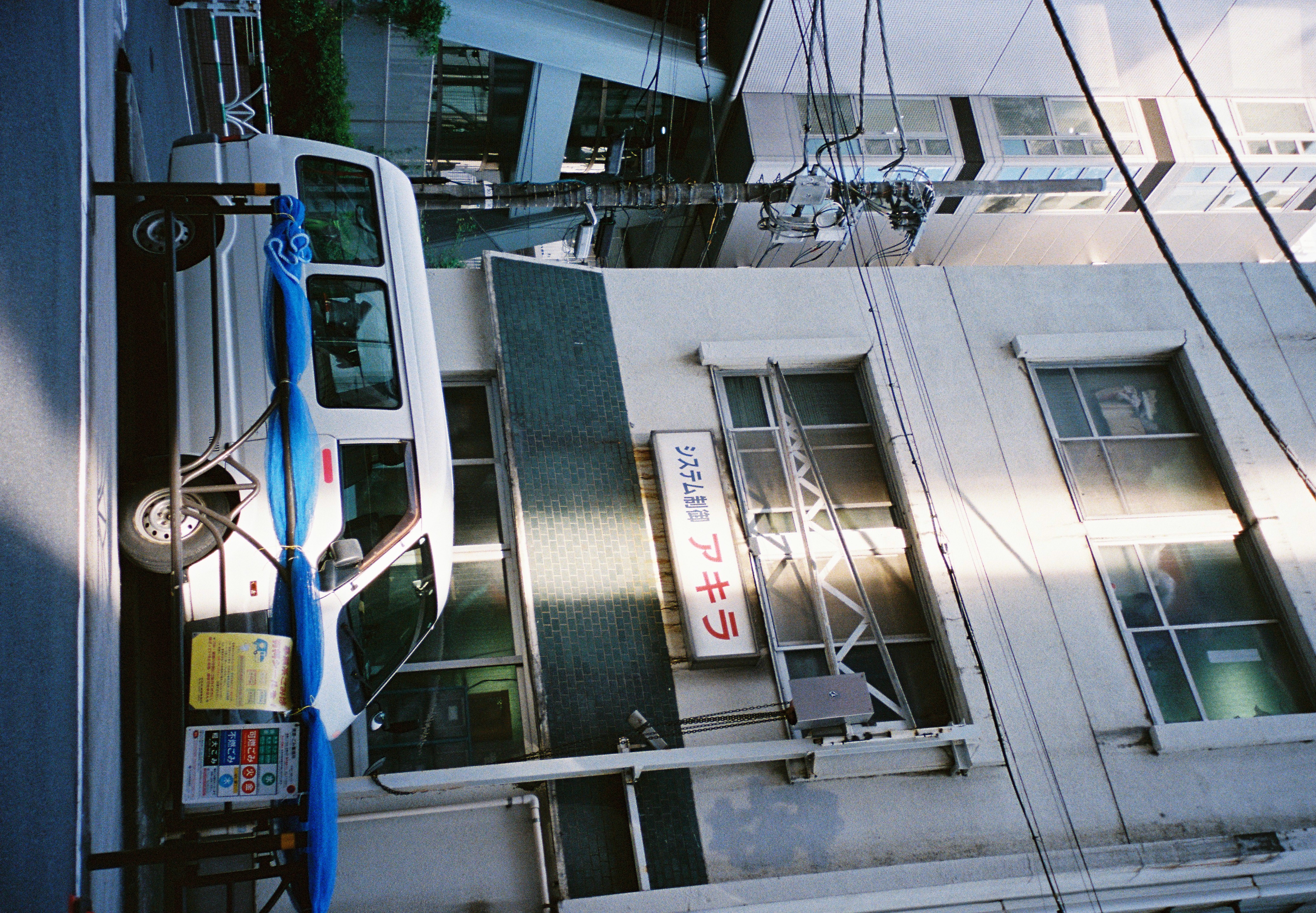 A white van with cargo wrapped in blue tarps sits parked beneath a signboard combining Japanese text with red katakana, above a green-tiled band.