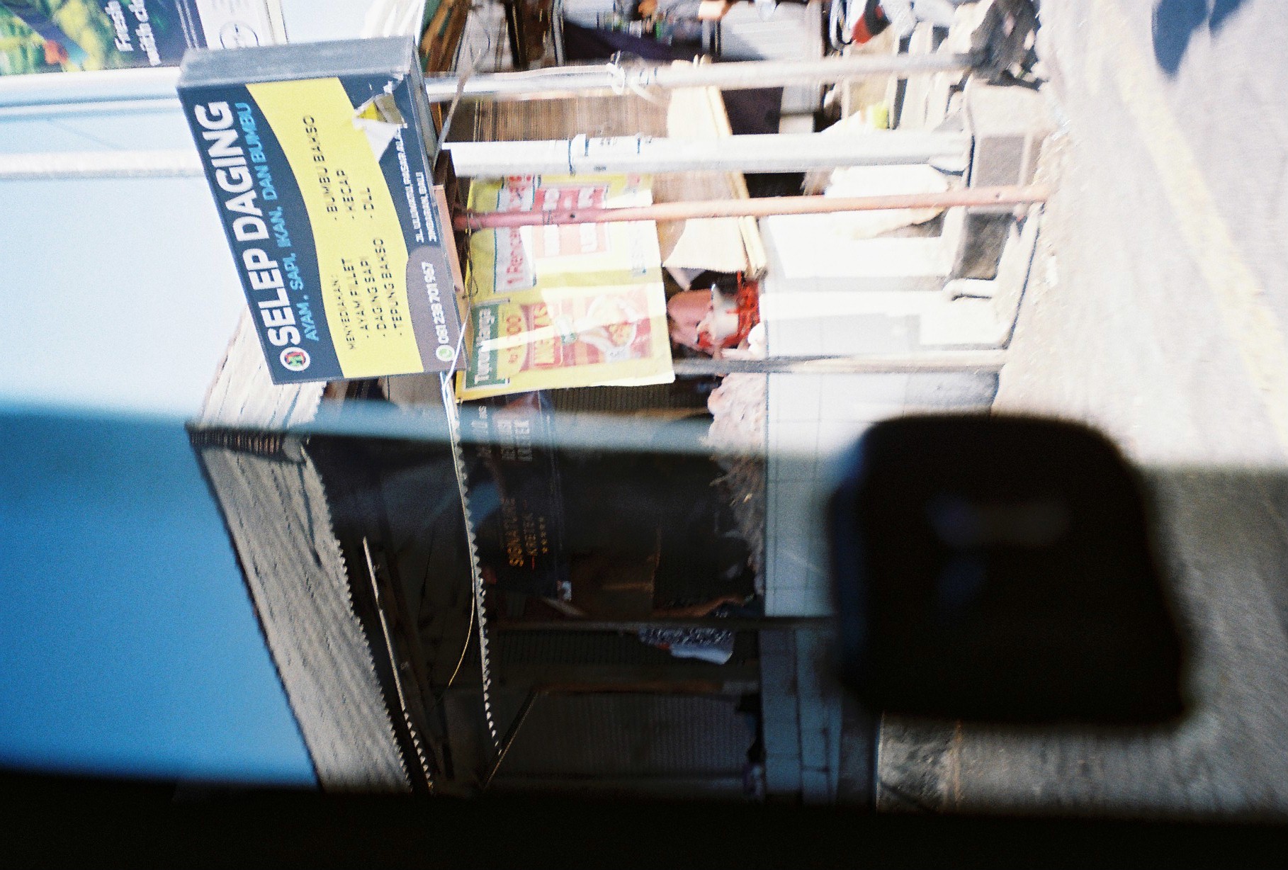 A tall sign reading “SELEP DAGING” stands above a small roadside stall, with dark blurred forms obscuring much of the left side.
