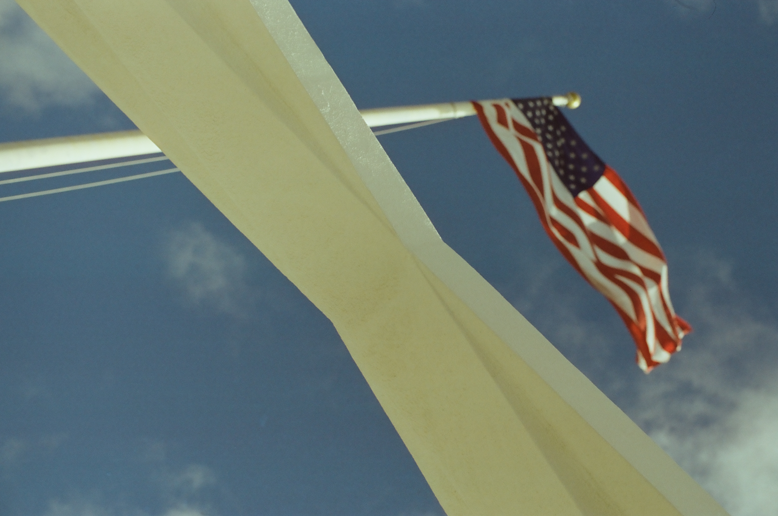 A softly focused American flag crosses between angular white beams beneath a deep blue cloudless opening.