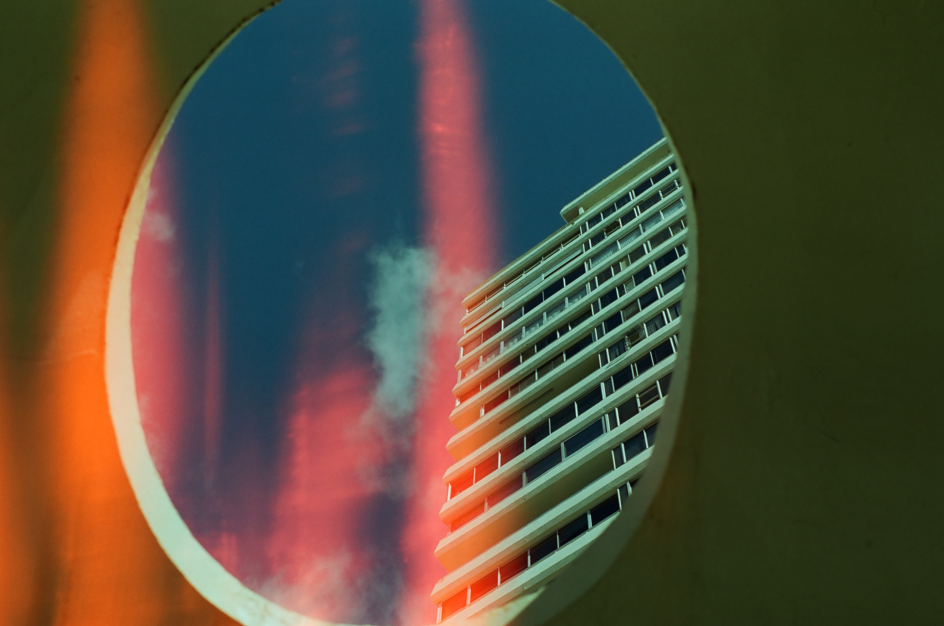 An oval concrete cutout frames a white high-rise's angled balconies beneath a streak of red light leaking across a deep blue sky.