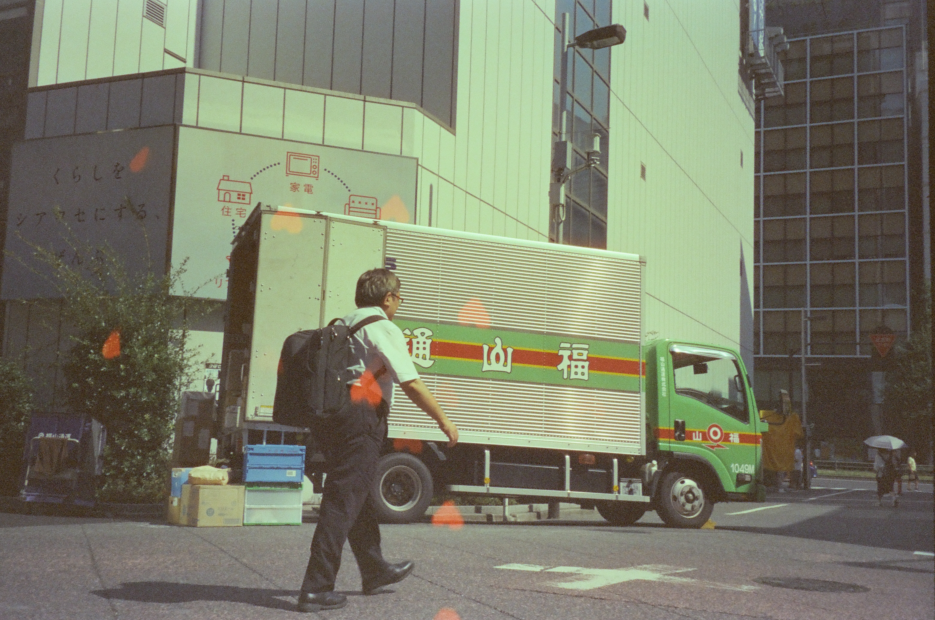 A man with a backpack walks past a green delivery truck marked with Japanese characters and the number '1049M,' with orange heart shapes scattered nearby.