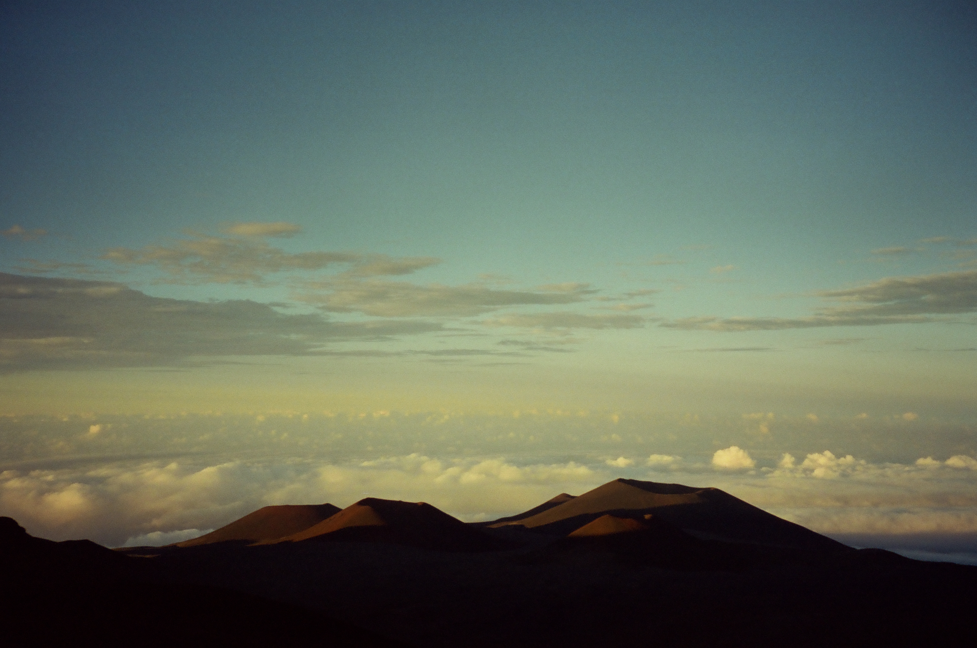 Layered dark ridges stretch above bright distant clouds beneath a green-blue evening sky.