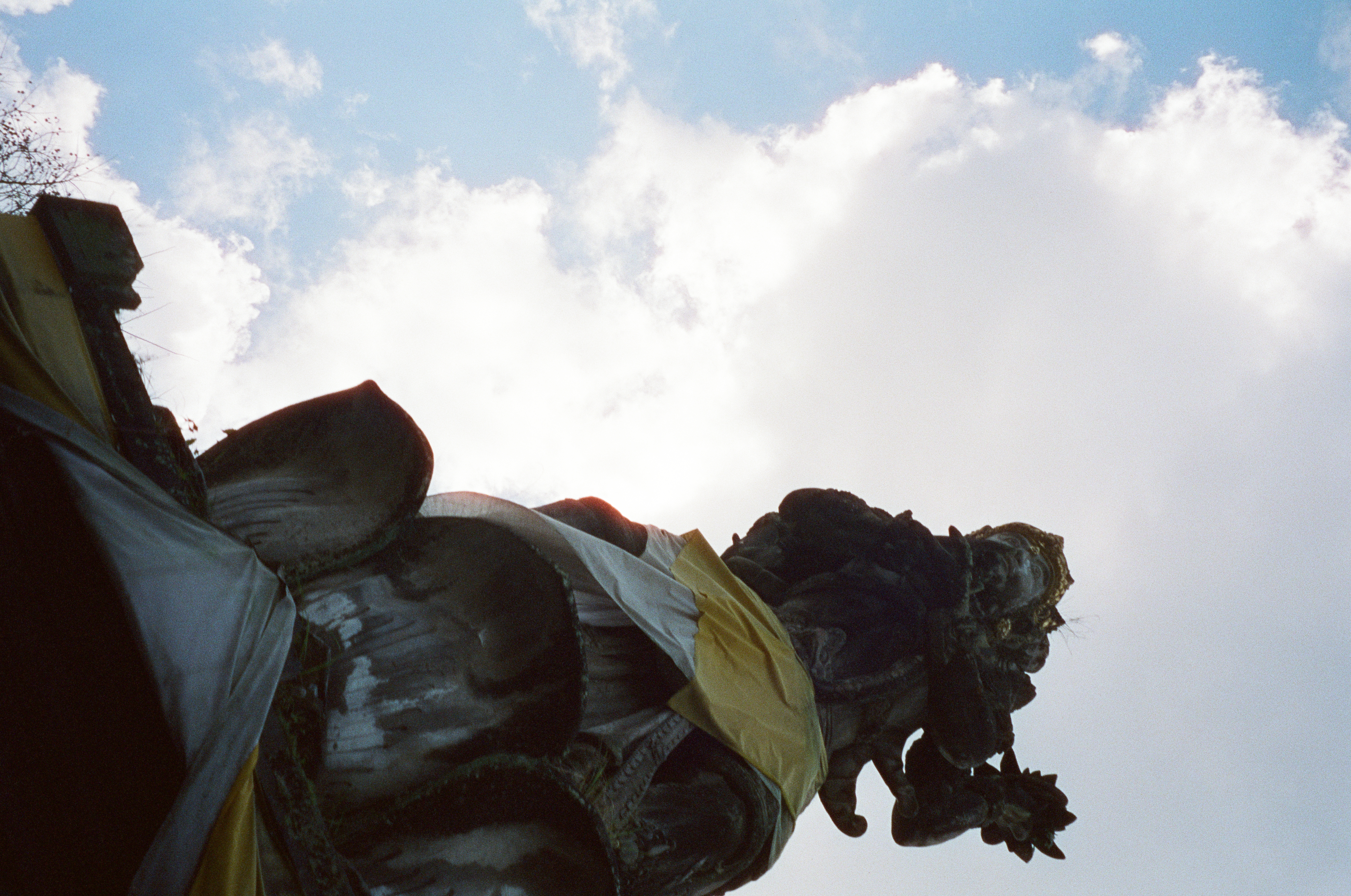 A dark statue wrapped in yellow and white cloth fills the right side of a low-angle view against bright clouds and blue sky.