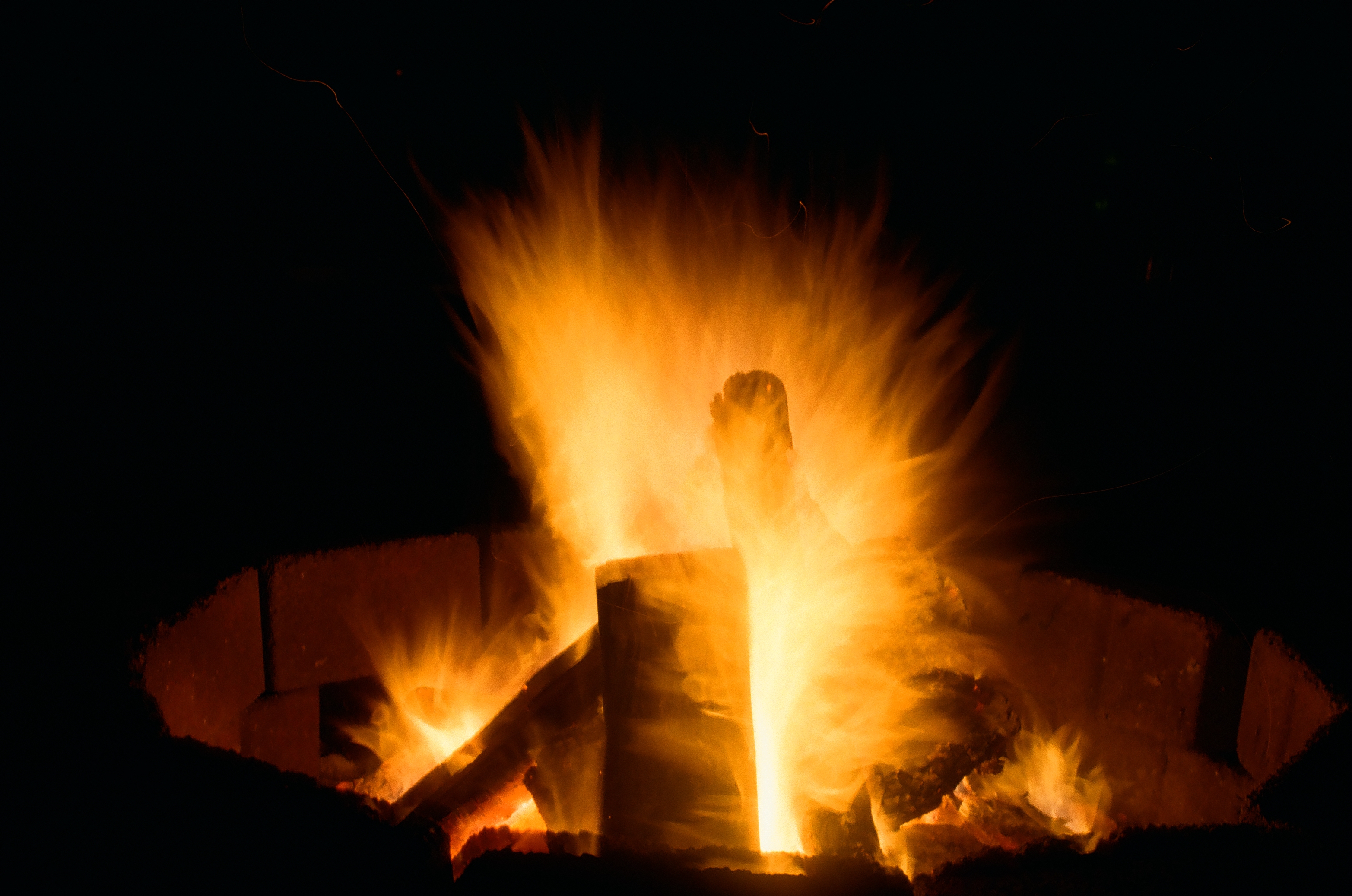 Tall orange flames rise from a circular fire pit at night, with a dark upright shape standing at the center of the blaze.