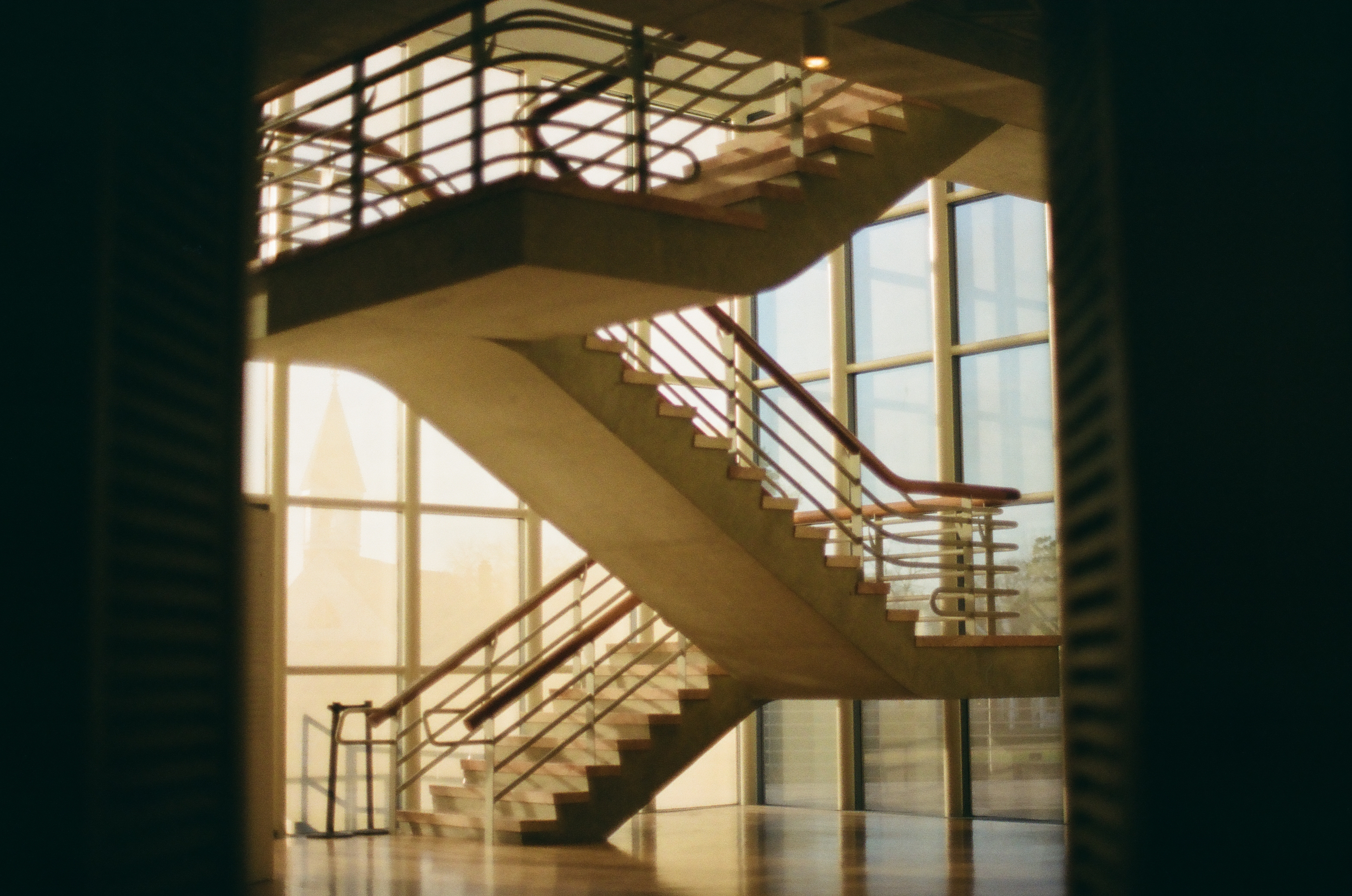 A switchback staircase with thin metal balusters and wooden handrails stands before a tall glass wall lit by warm daylight.