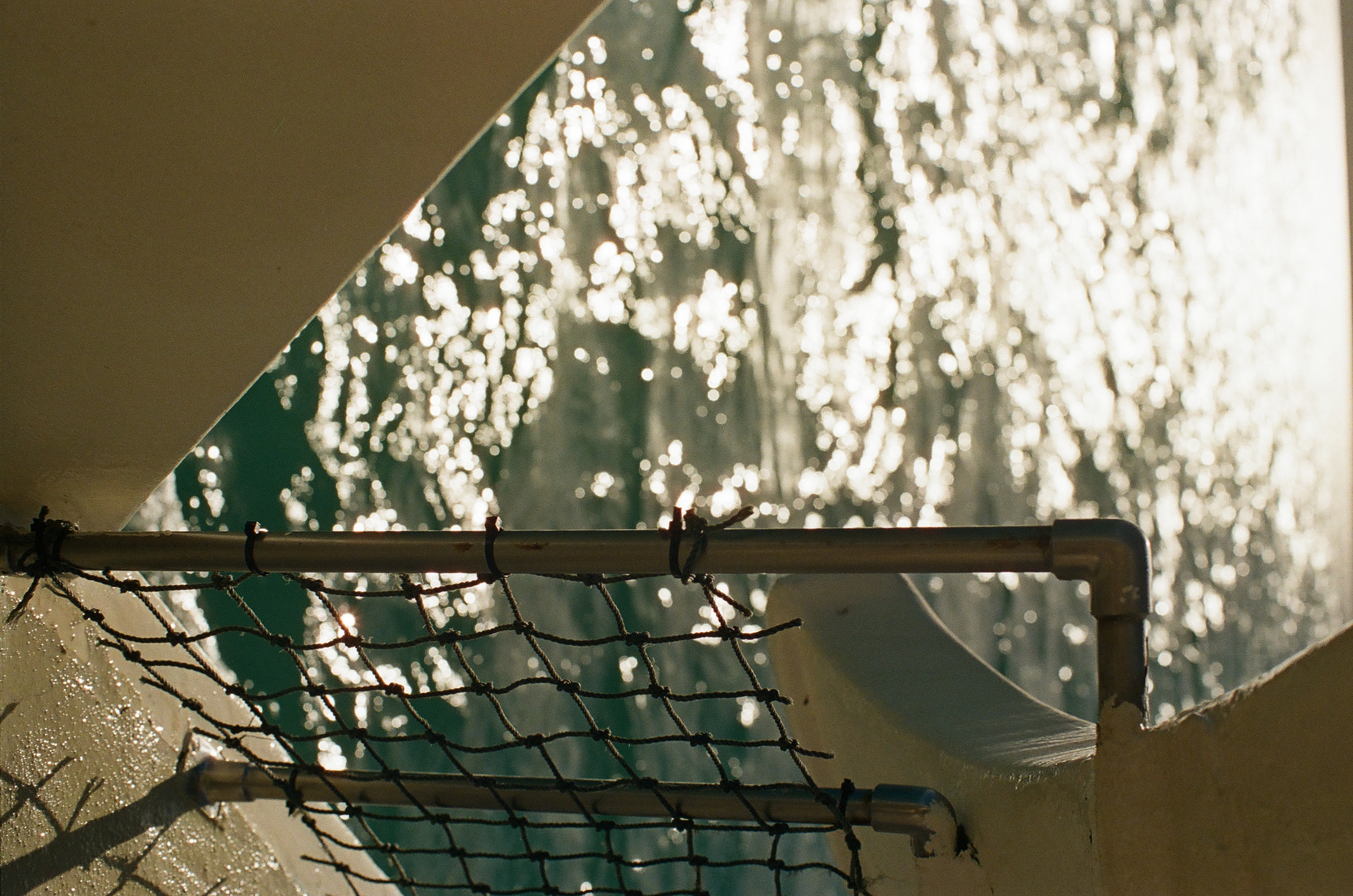 Sunlight scatters into bright bokeh across choppy water behind a metal rail wrapped in knotted rope netting on the boat's side.
