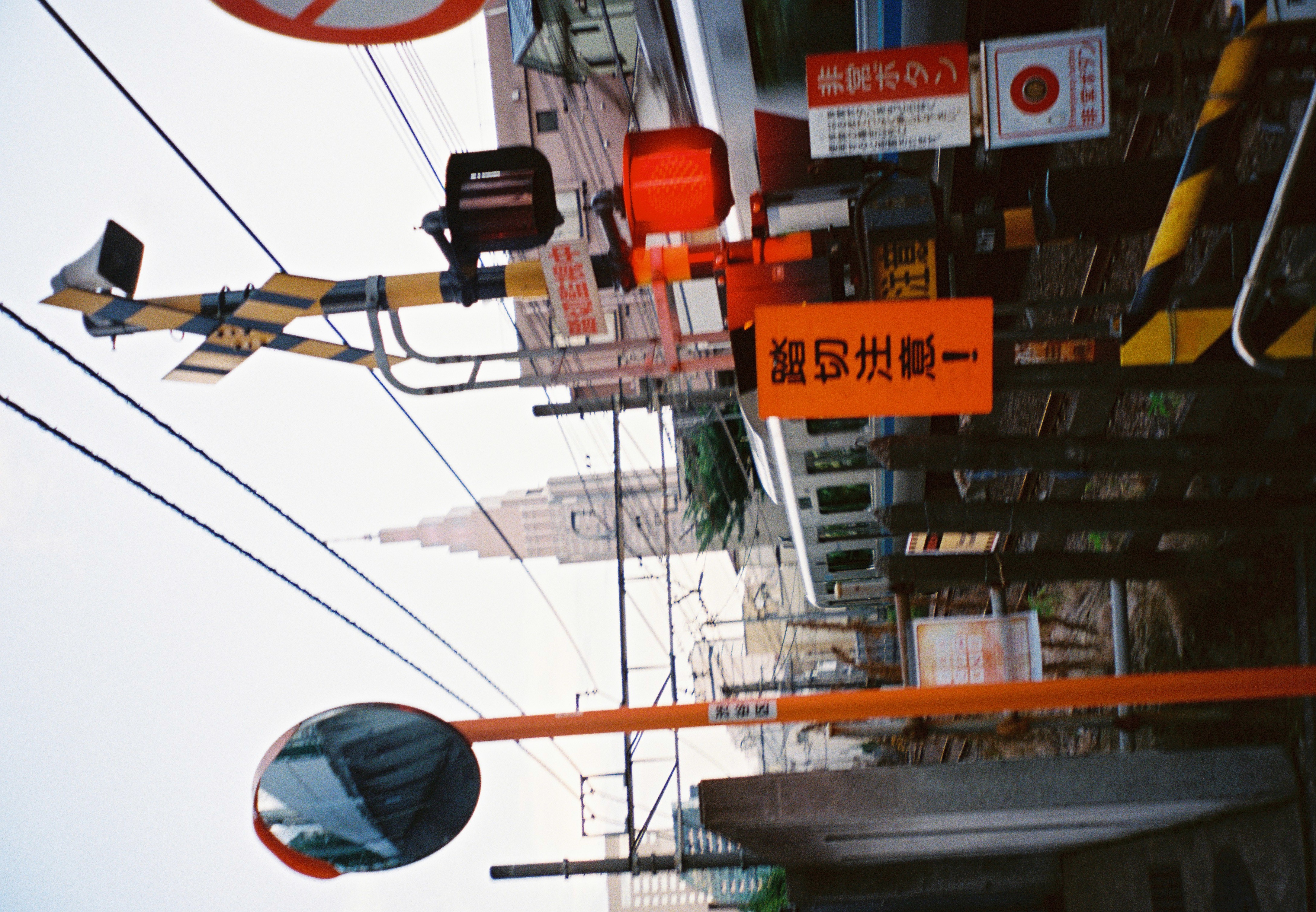 An orange-and-black railway crossing stands among overhead cables, warning signs, mirrors, and nearby buildings.