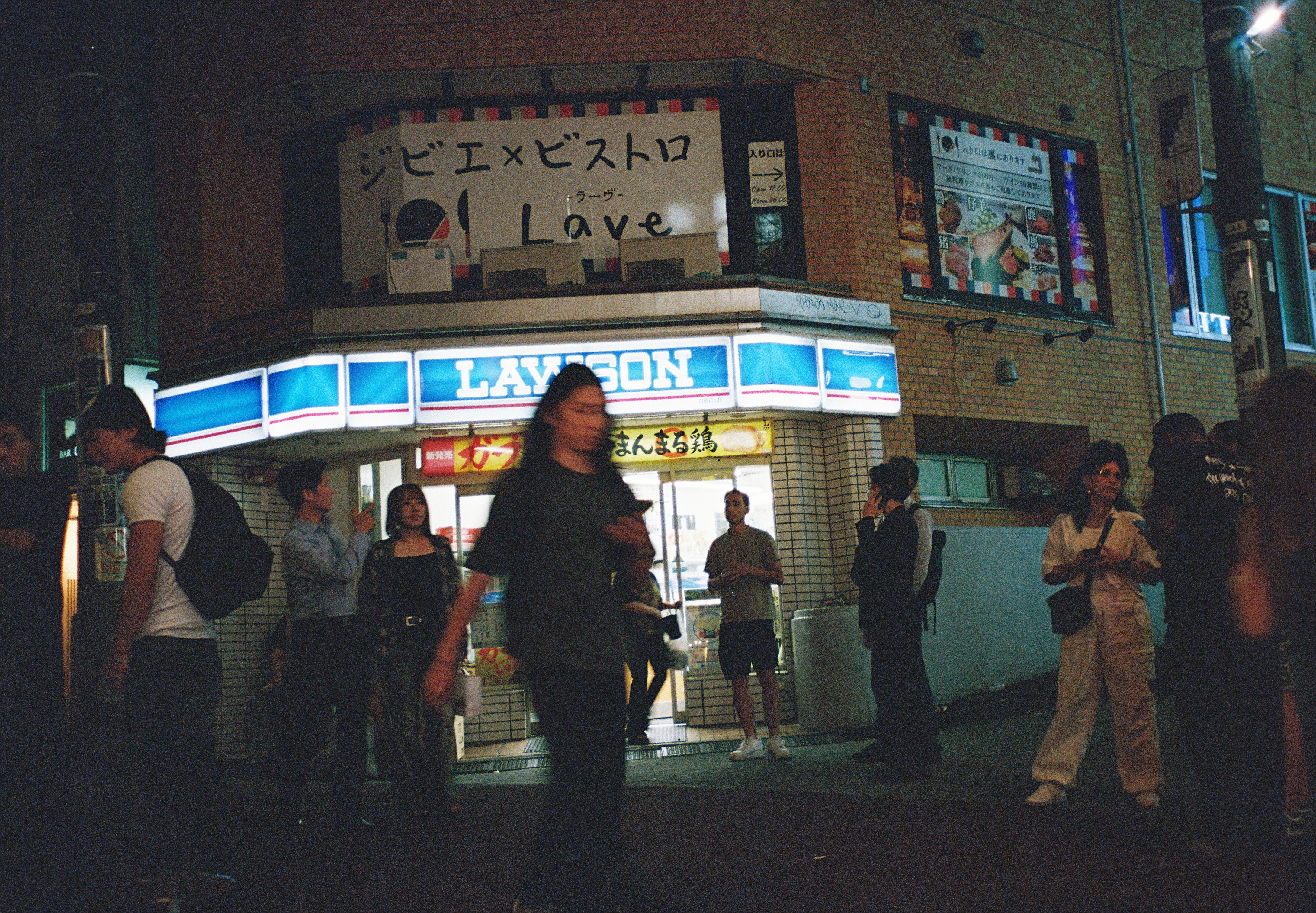 A motion-blurred pedestrian crosses in front of a brightly lit Lawson storefront while several other people stand near its entrance.
