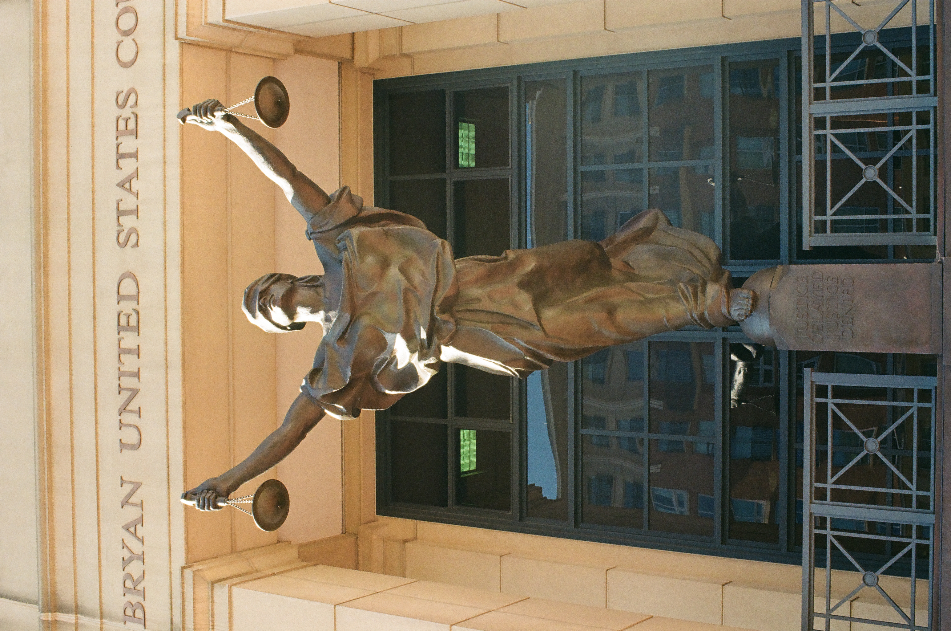 A bronze female figure raises scales and a sword before a pale courthouse facade and dark entrance.