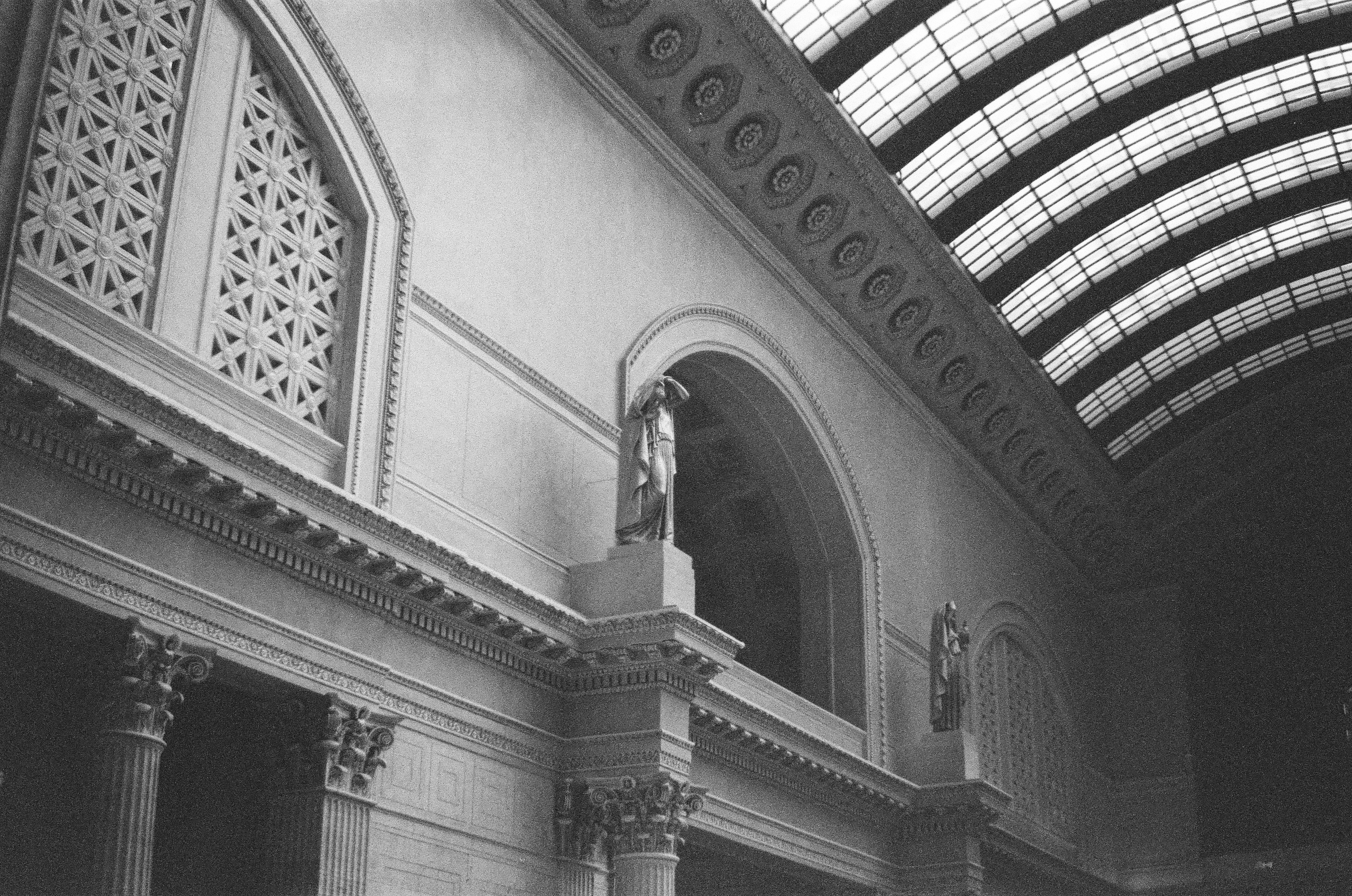 An arched interior wall, decorative lattice, standing statue, and curved glass ceiling rise through soft monochrome light.