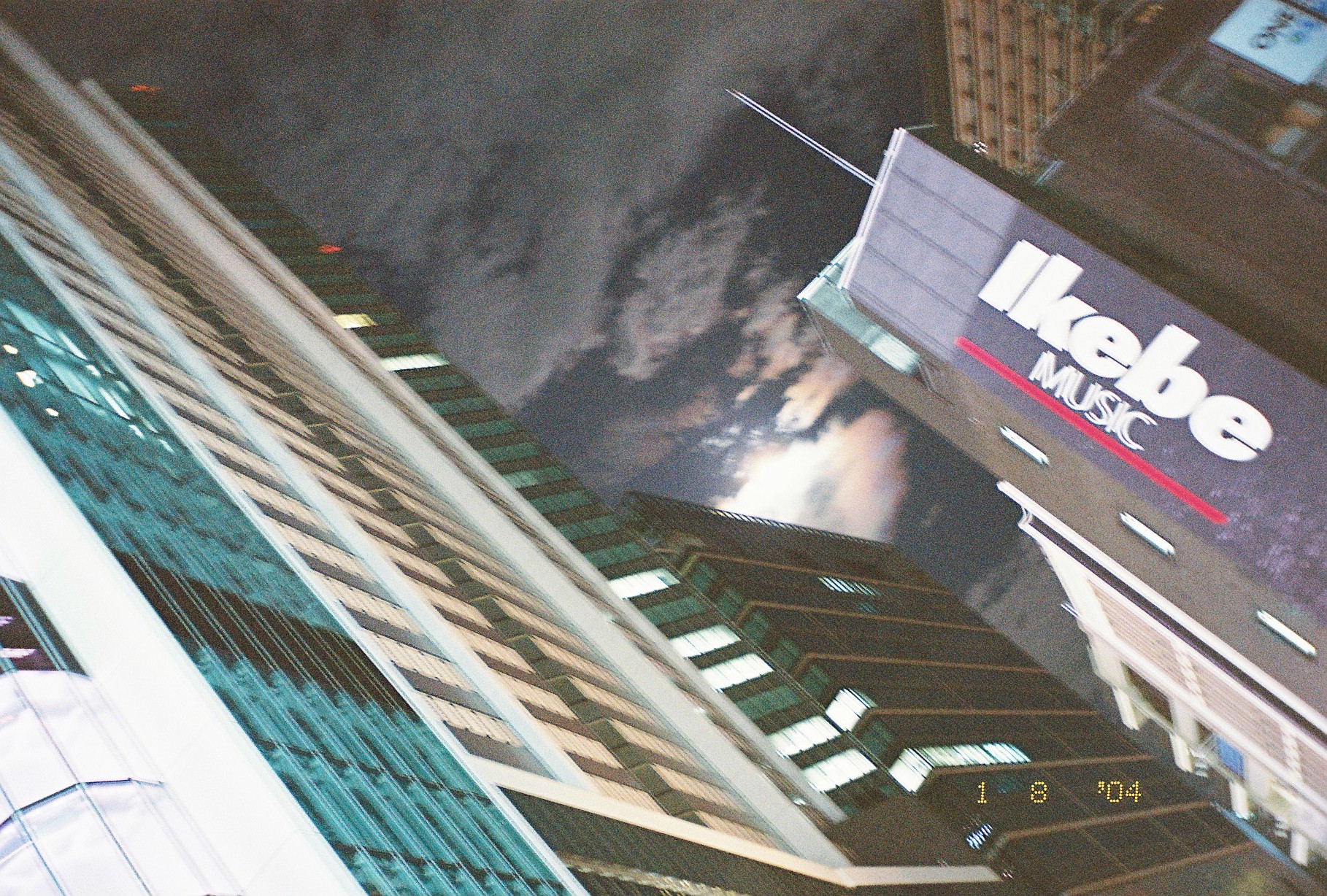 A glass high-rise tilts across the frame beside a sign reading Ikebe Music, with the moon glowing through thick clouds above.