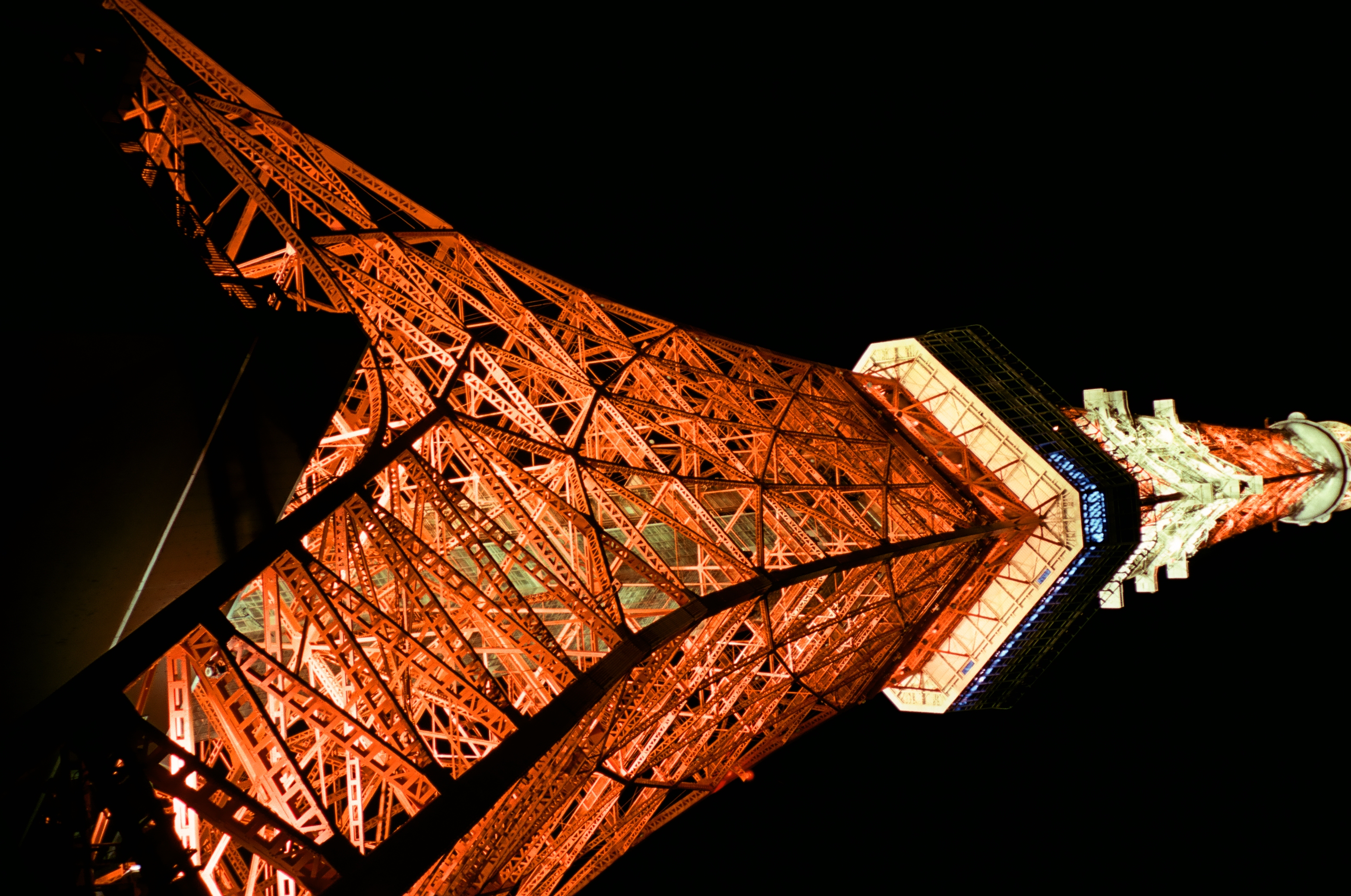 Steeply angled orange girders converge upward toward the white-lit underside of a platform against a black sky.