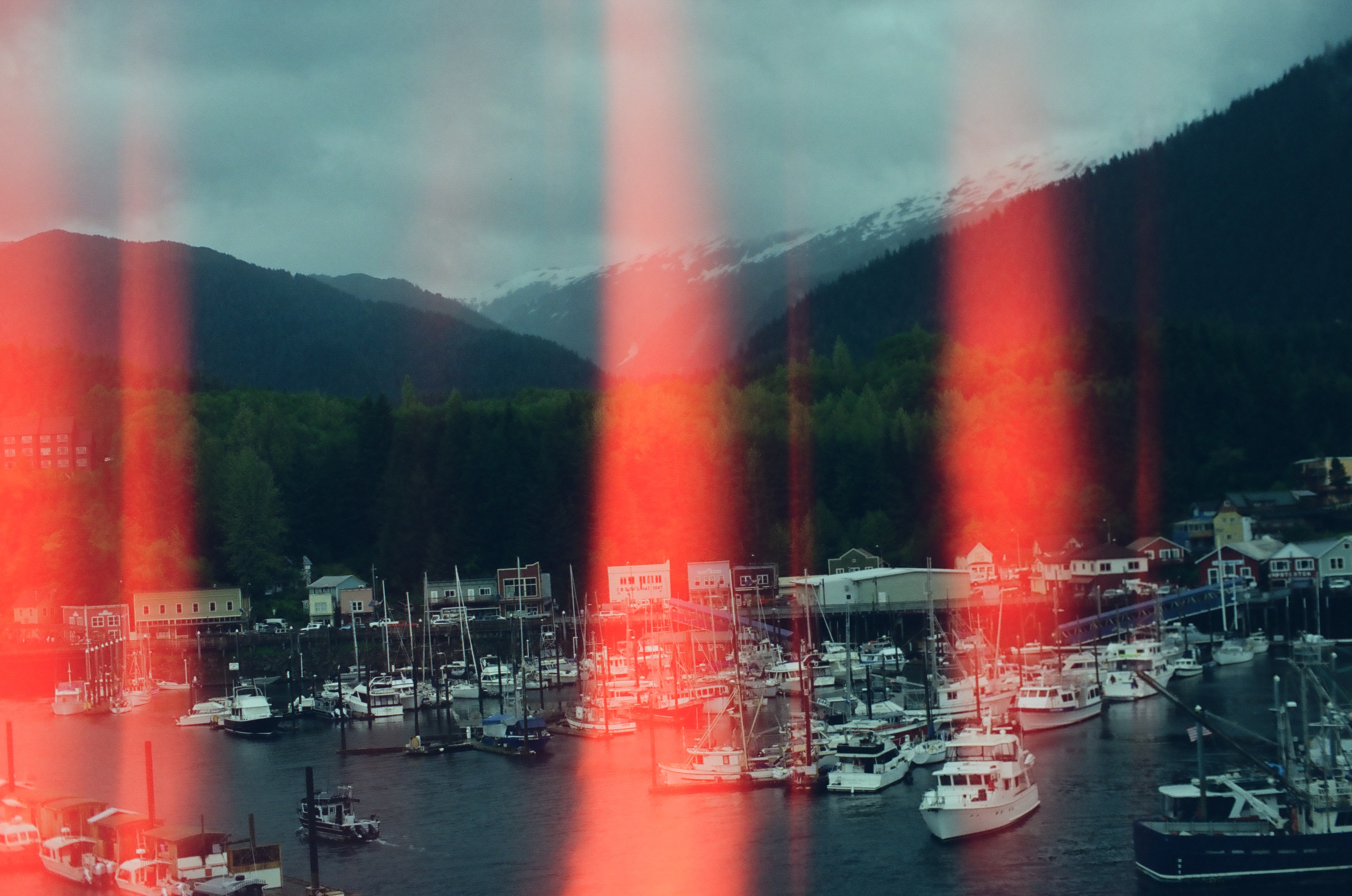 Vertical red light leaks streak across a view of a marina full of moored boats beneath snow-capped mountains and forest.