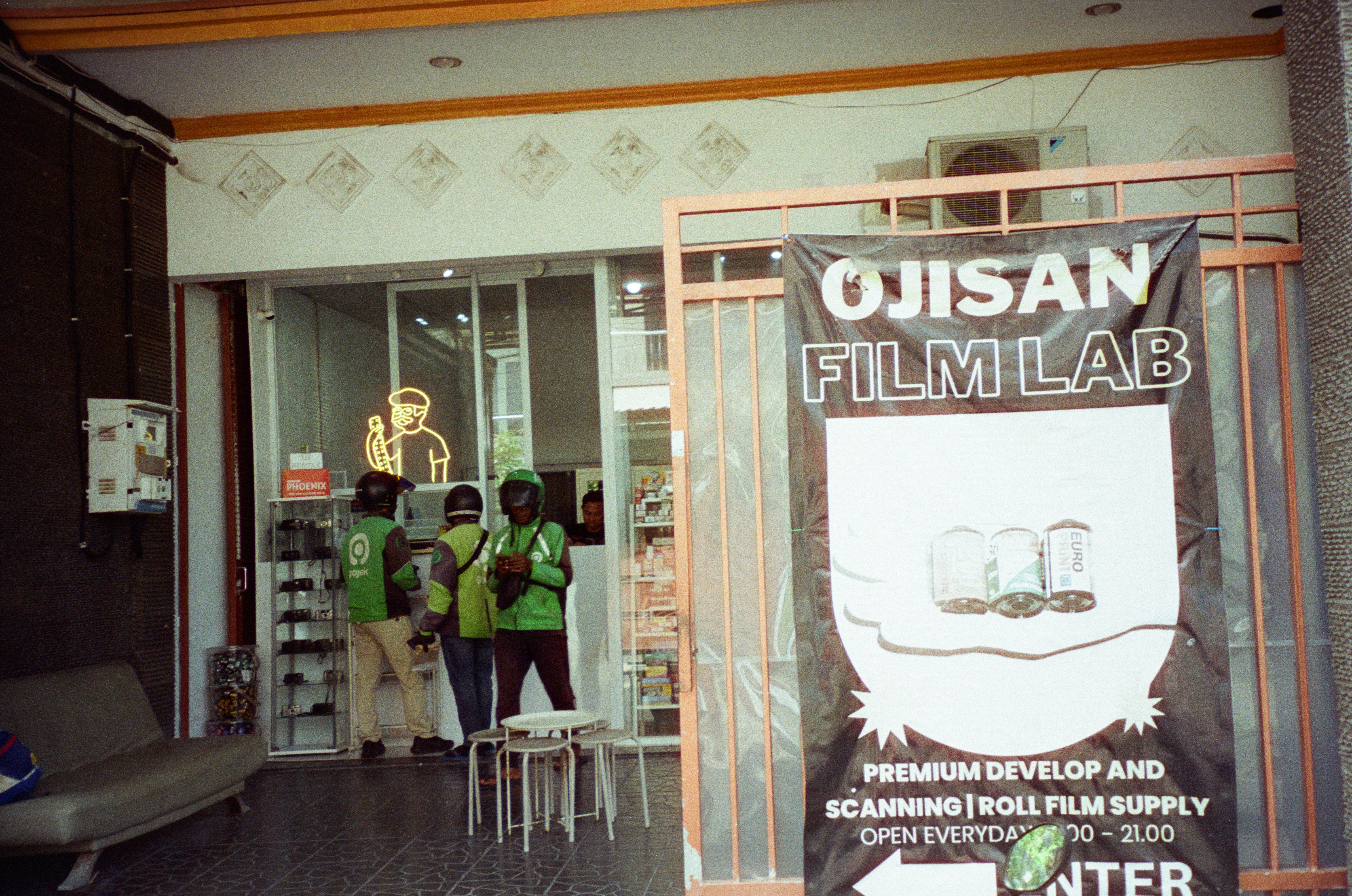 Three helmeted people in green jackets stand at a shop counter beside a tall black banner reading OJISAN FILM LAB, premium develop and scanning, roll film supply.