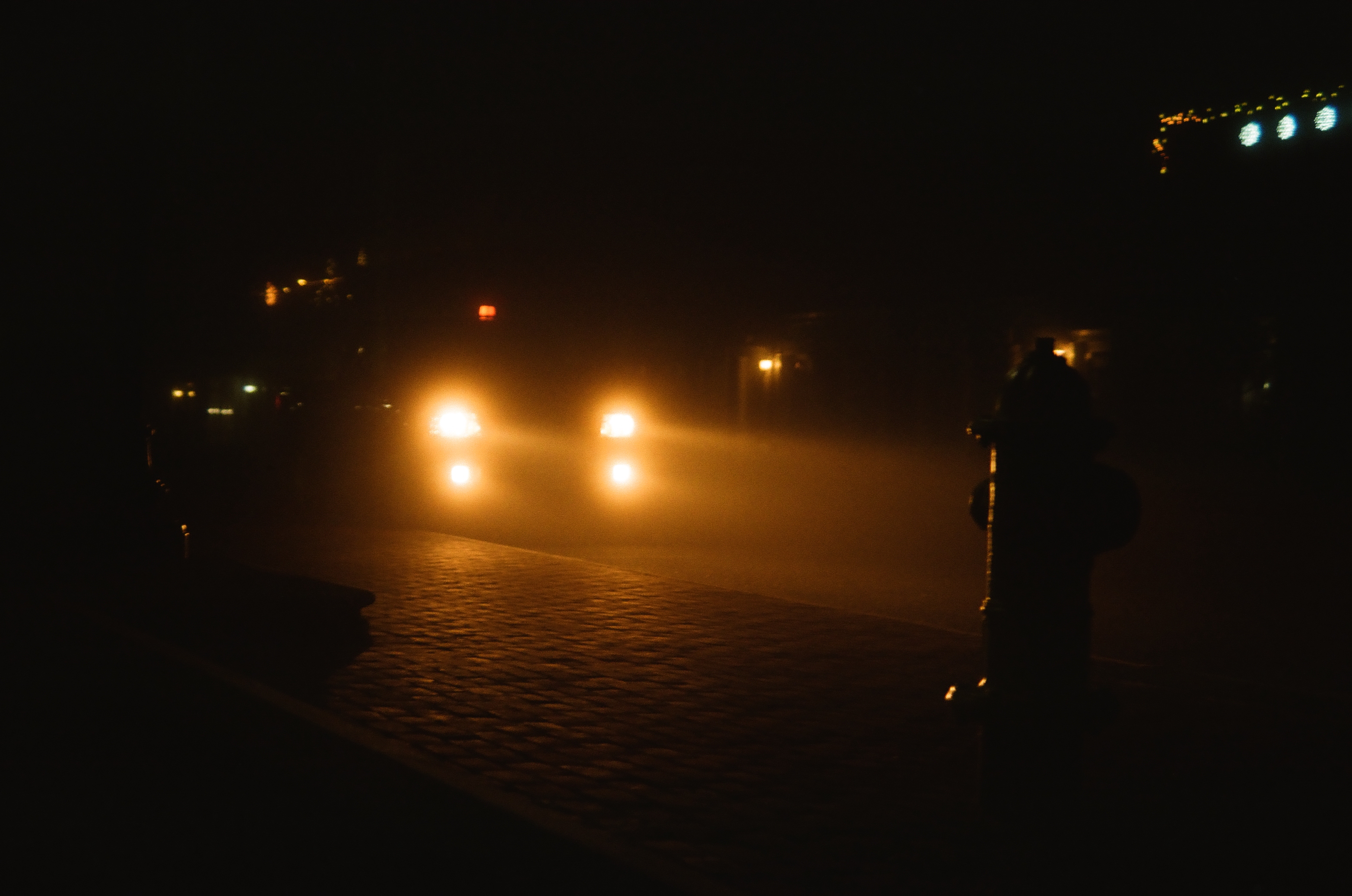 Two vehicle headlights glow through dense fog on a brick-paved street, with a fire hydrant silhouetted at the right edge.