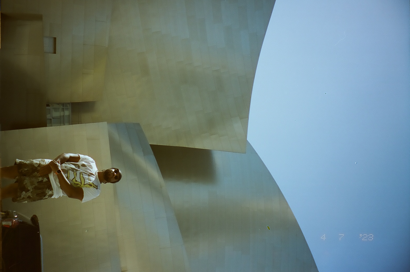 A person stands small beneath a towering curved metal facade in a narrow vertical composition.