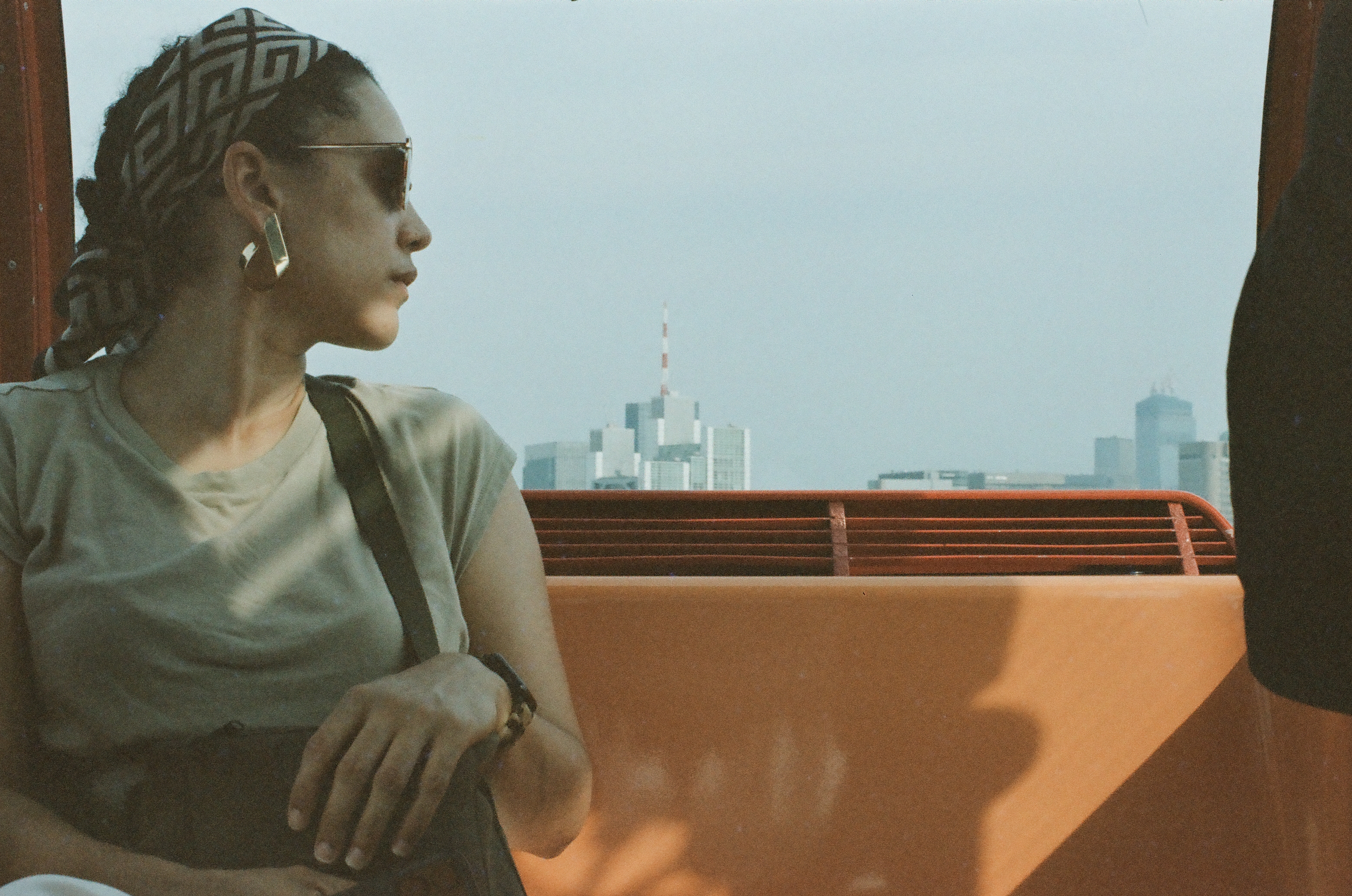 The same woman turns to look out at a hazy skyline of high-rises and a slender lattice tower beyond the gondola's orange rail.