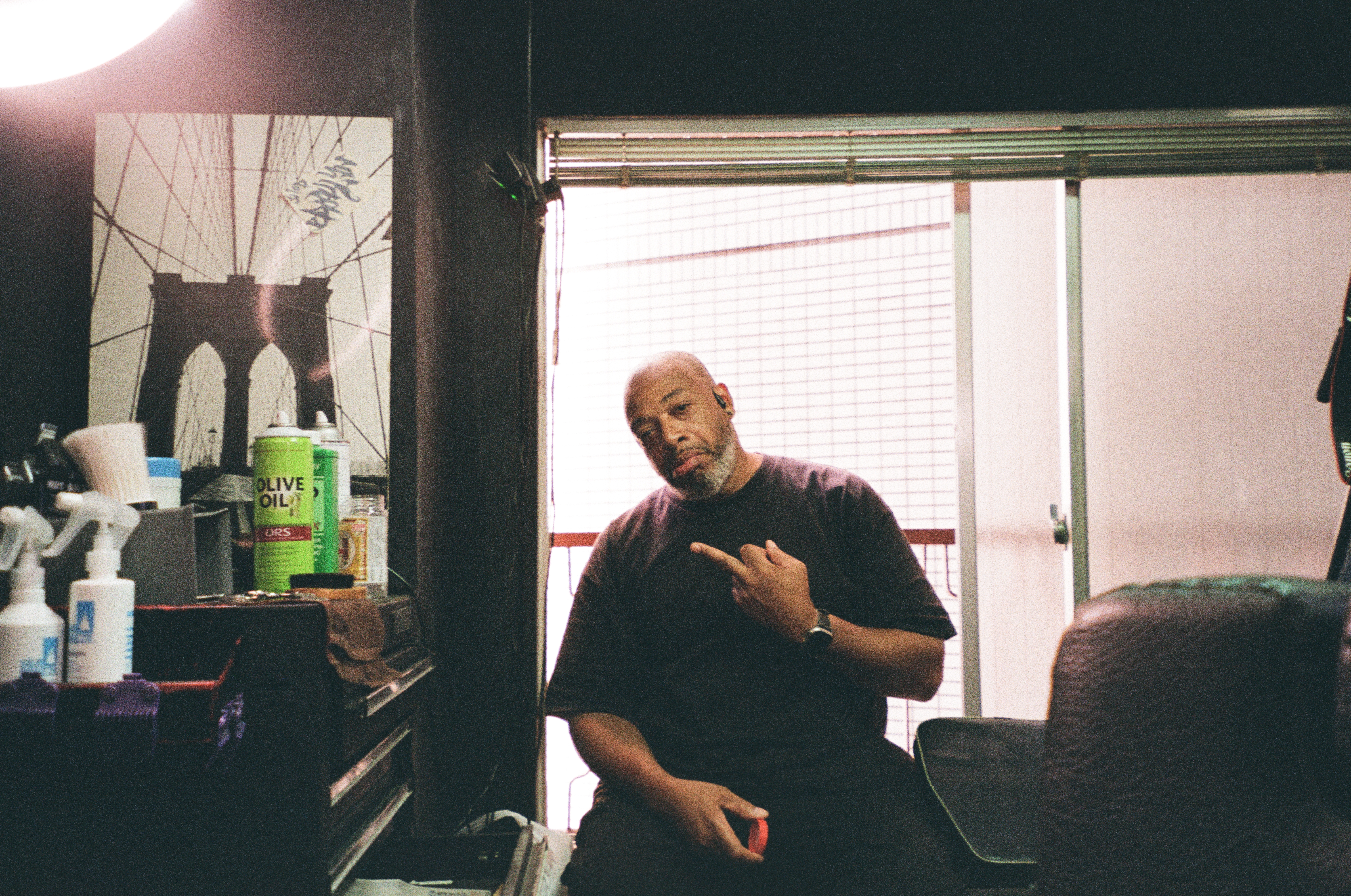 A seated man in a dark T-shirt raises two fingers in front of a bright window with blinds gathered at the top, near spray bottles and a green can.