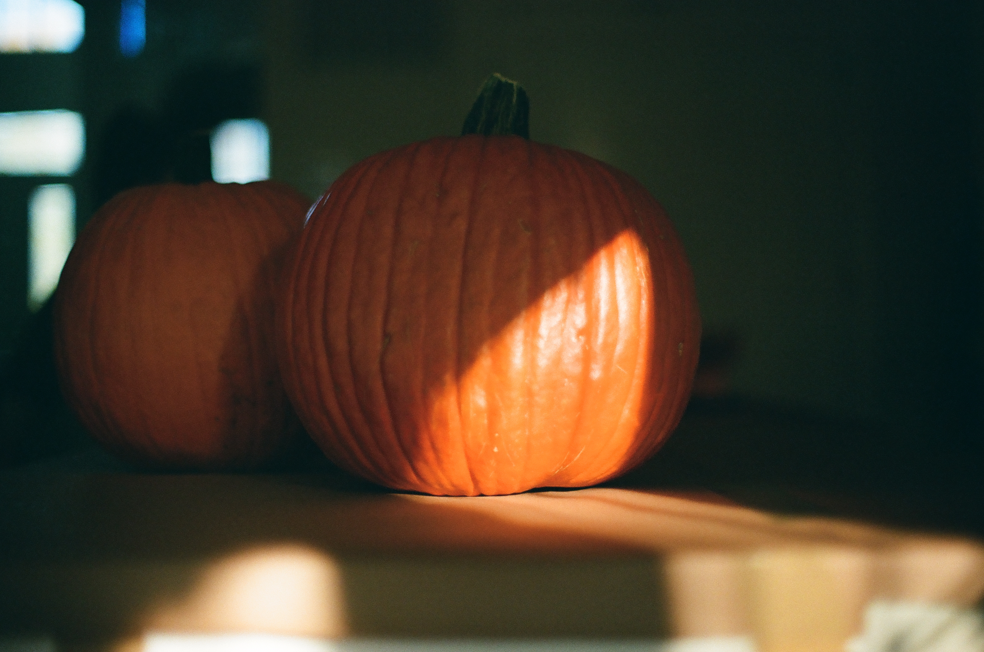 Two orange pumpkins rest on a dark ledge while a diagonal bar of sunlight warms the larger fruit.
