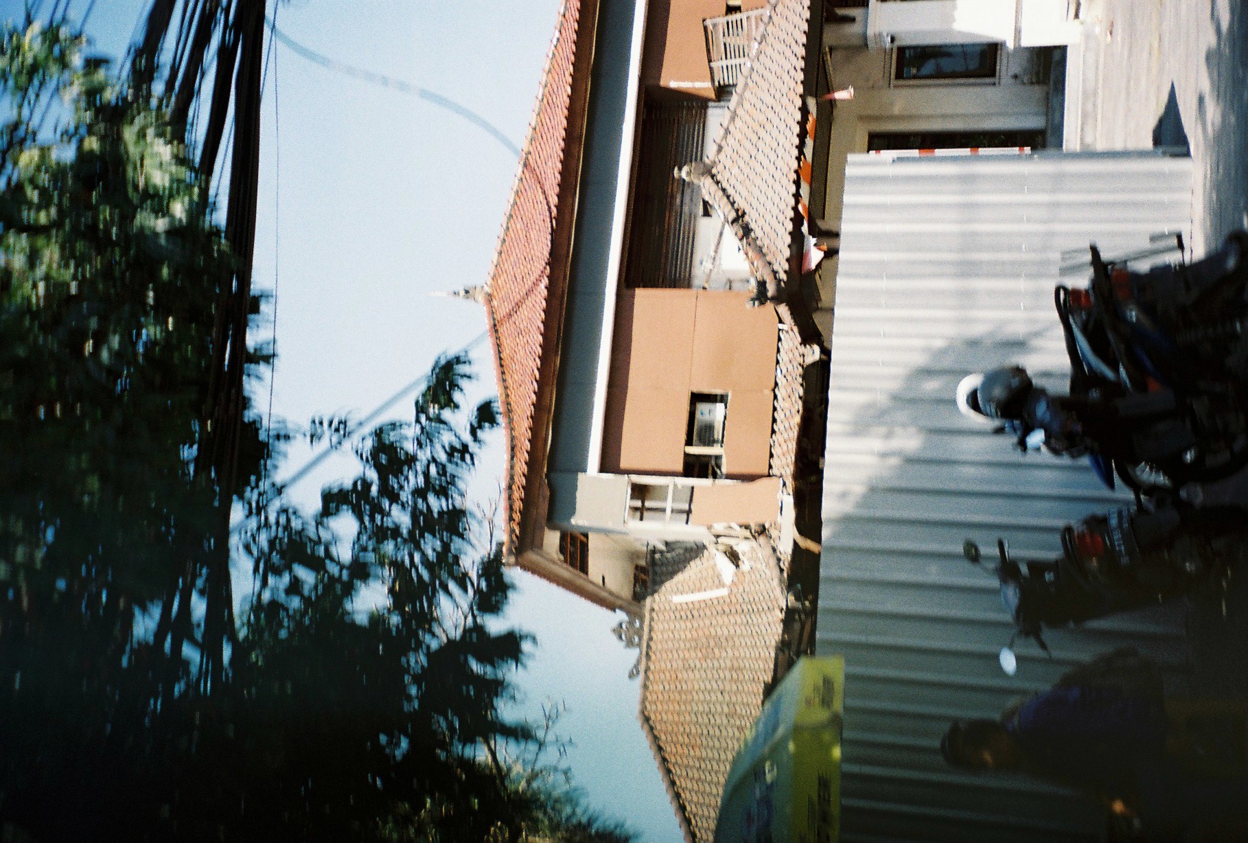 Scooters rest in shade beneath overhead wires and a pale building capped with reddish tile roofs.