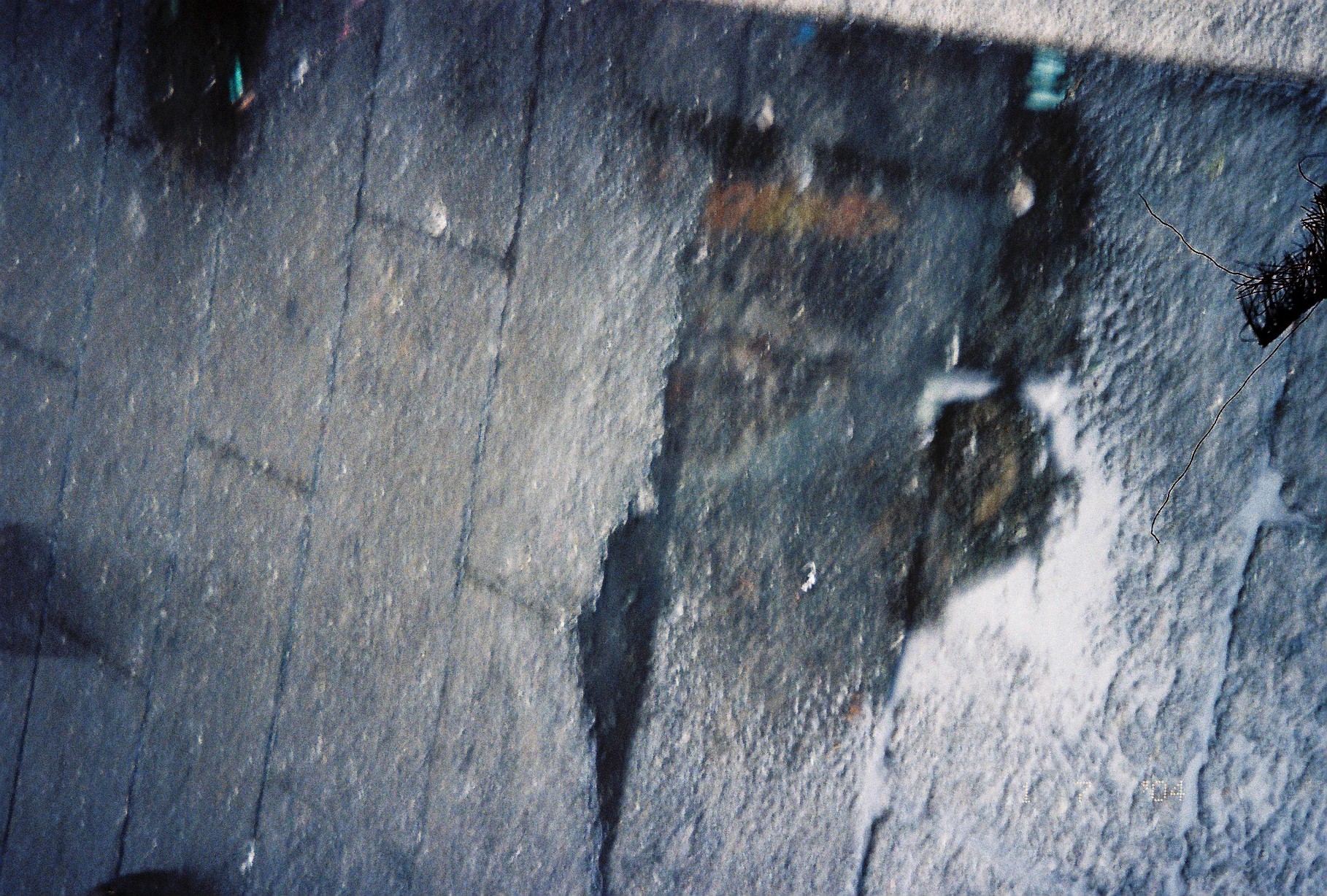 A rain puddle on cracked stone pavement holds a blurred, dark reflection of an indistinct hunched shape above pale sky.