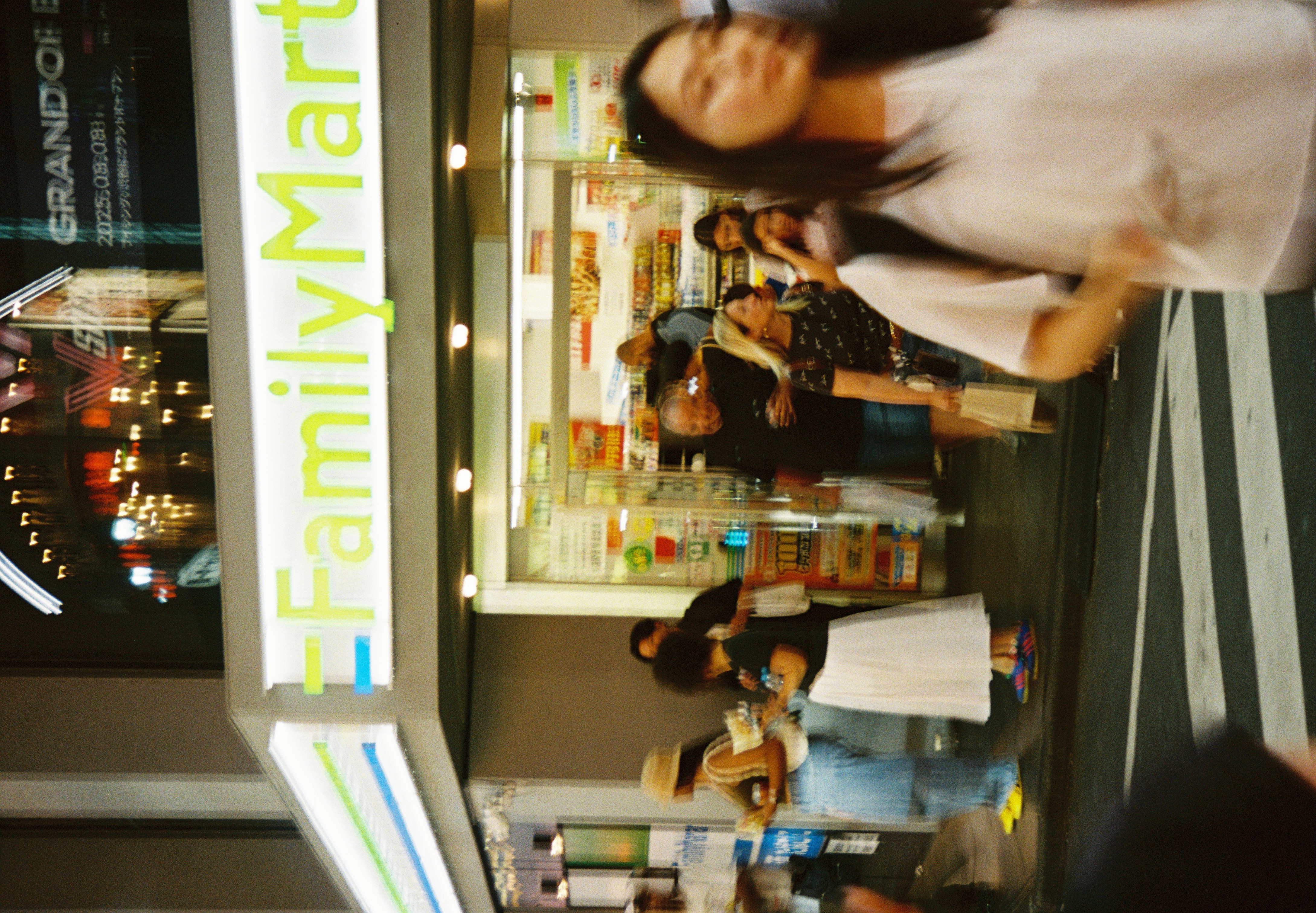 Several people stand and walk outside a convenience store lit by a green 'FamilyMart' sign, with a blurred figure crossing painted road stripes.