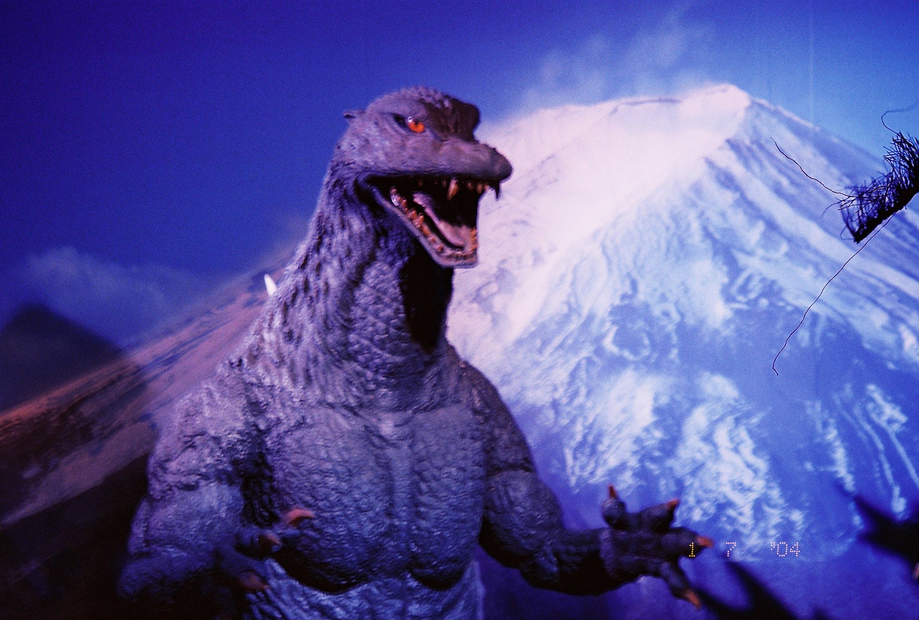A sculpted creature figure with lit orange eyes stands before an illuminated painted backdrop of a snow-covered mountain under deep blue lighting.