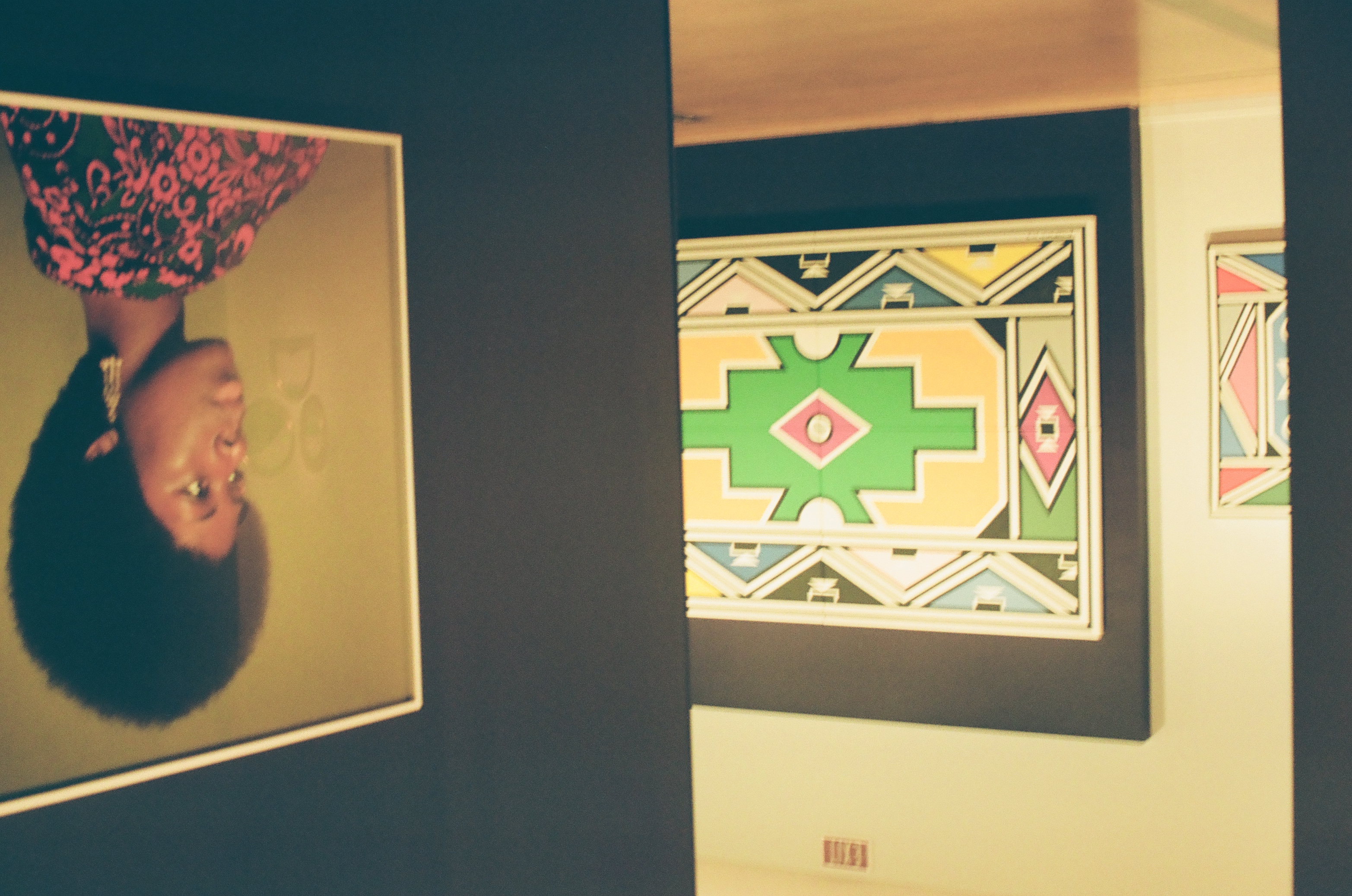 A gallery view with a large geometric painting in green, yellow, pink, and blue on a dark wall and a framed portrait of a woman with an afro.