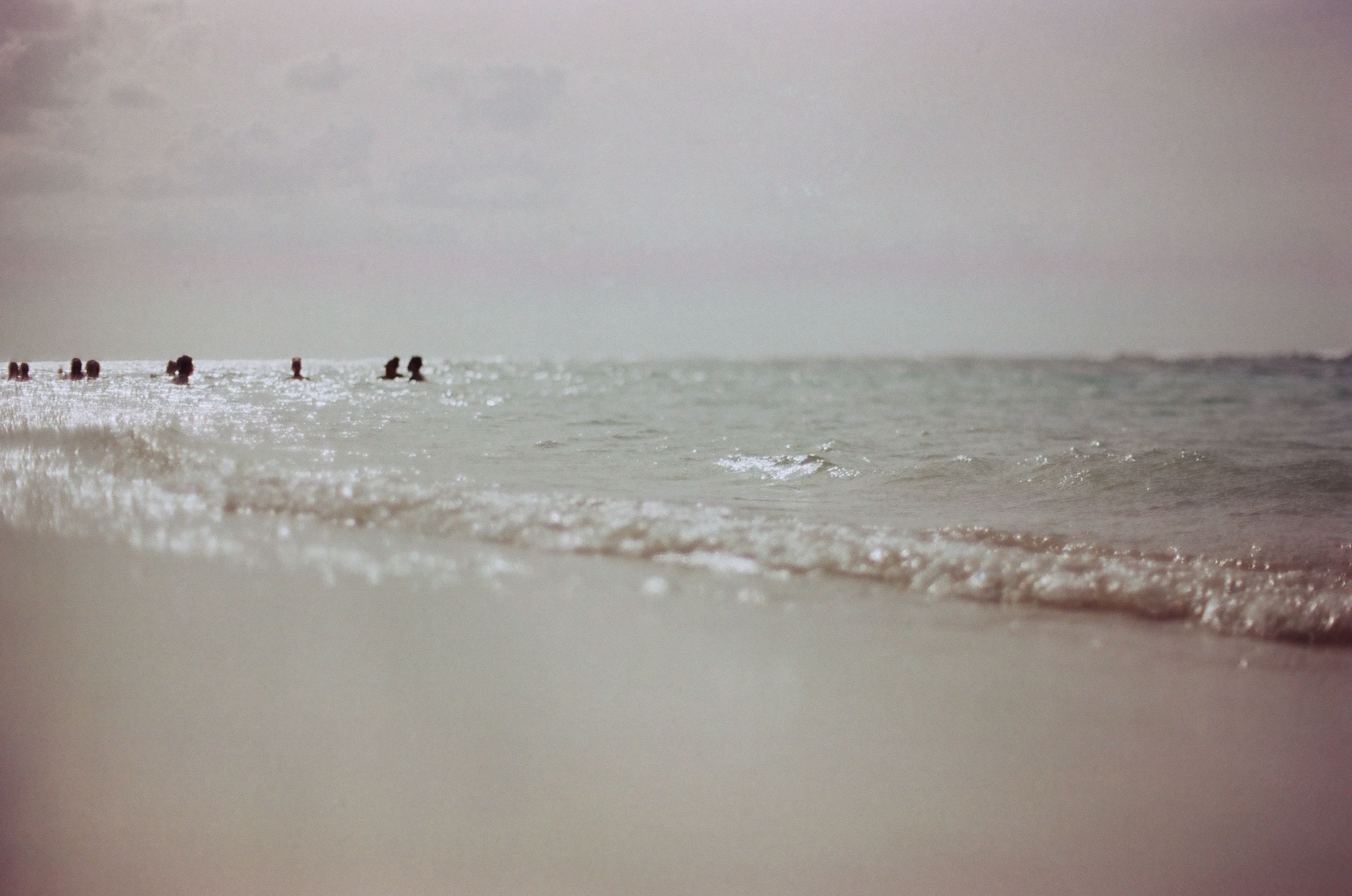 Low view across foamy shallow surf toward a scattered group of swimmers' heads above the water at left under a pale hazy sky.
