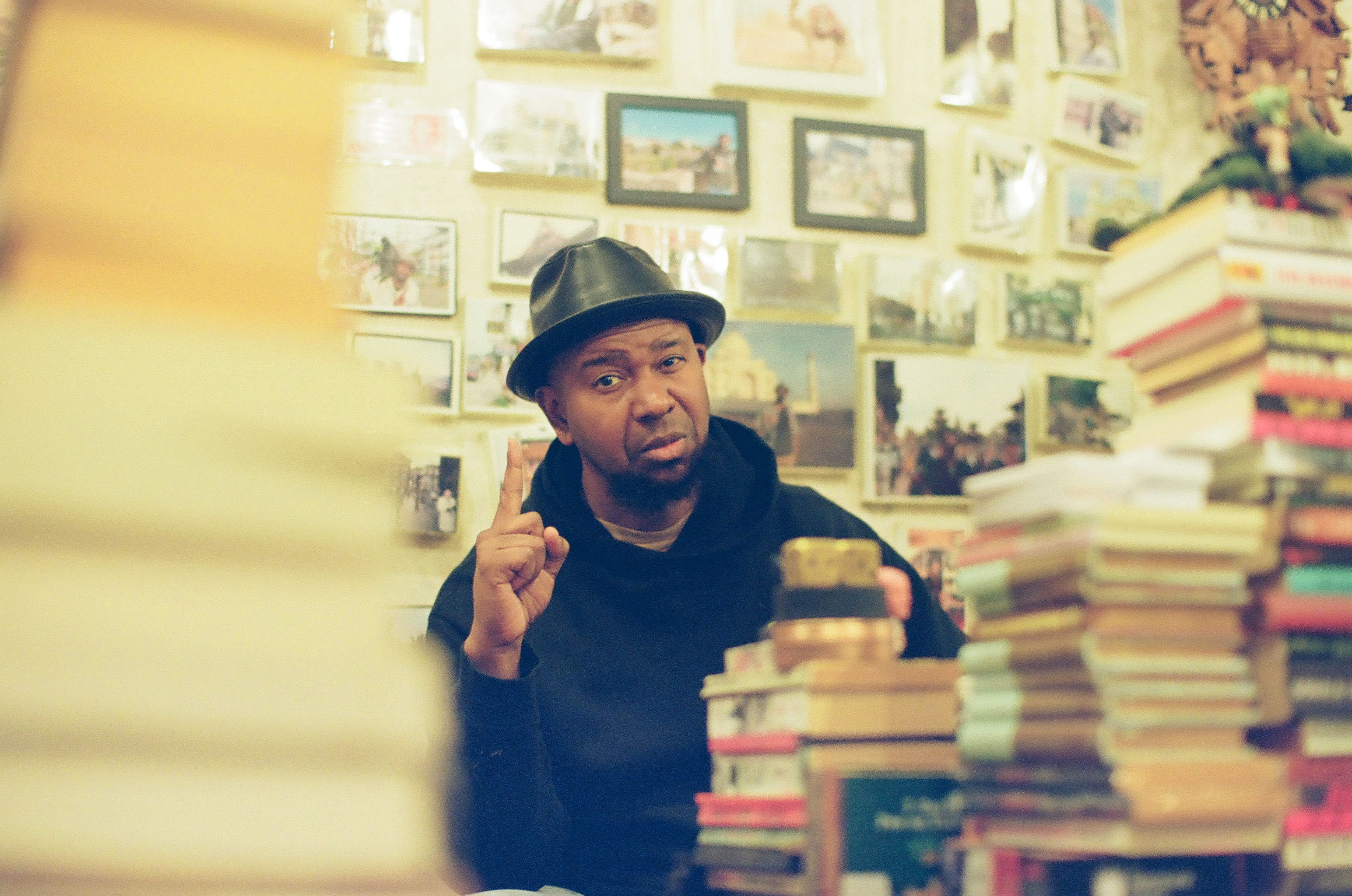 A seated man raises one index finger between a pale foreground blur, stacked books, and framed pictures.