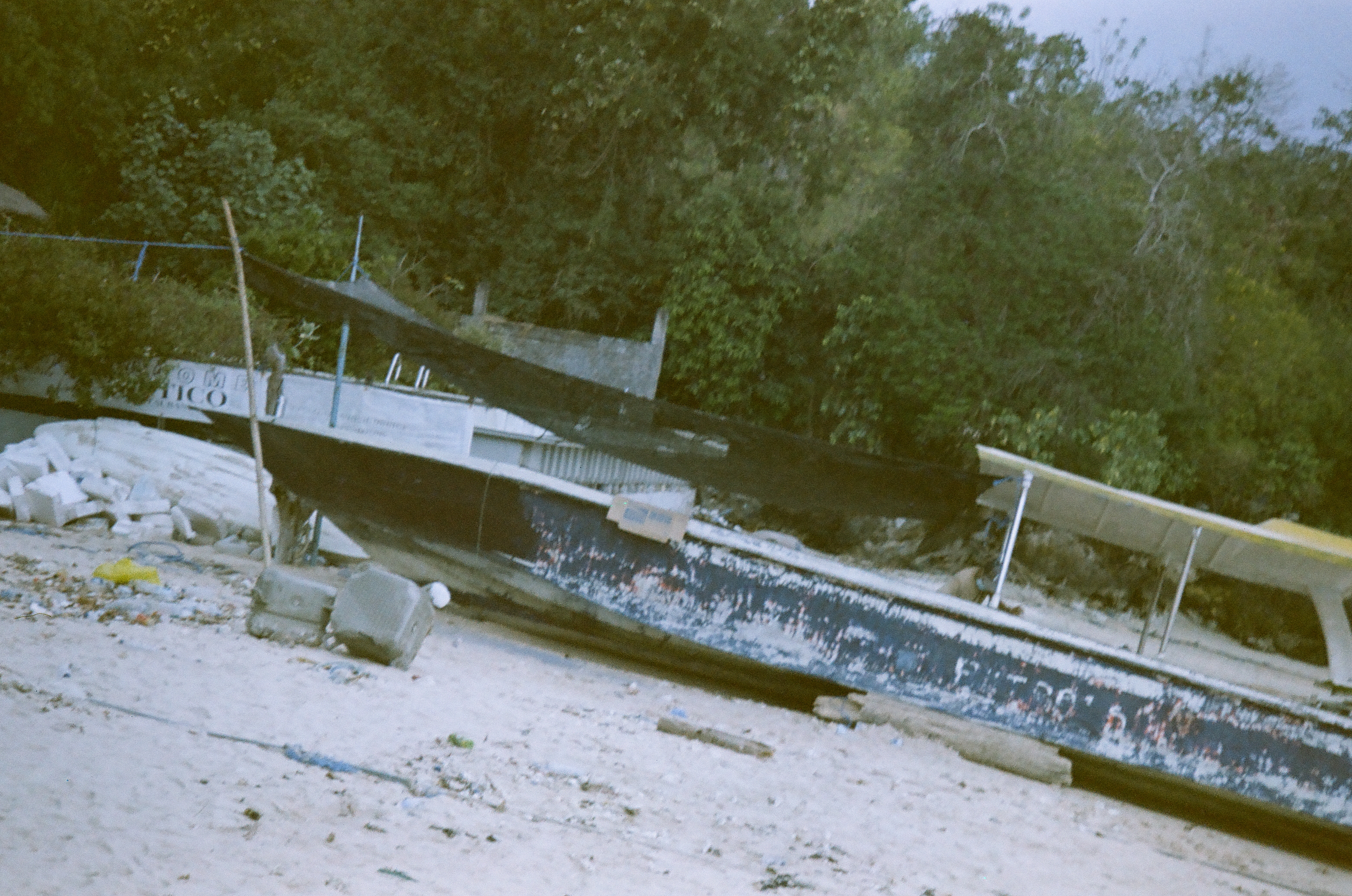 A long weathered boat rests on pale ground beneath dark material stretched between poles, with dense green trees filling the background.