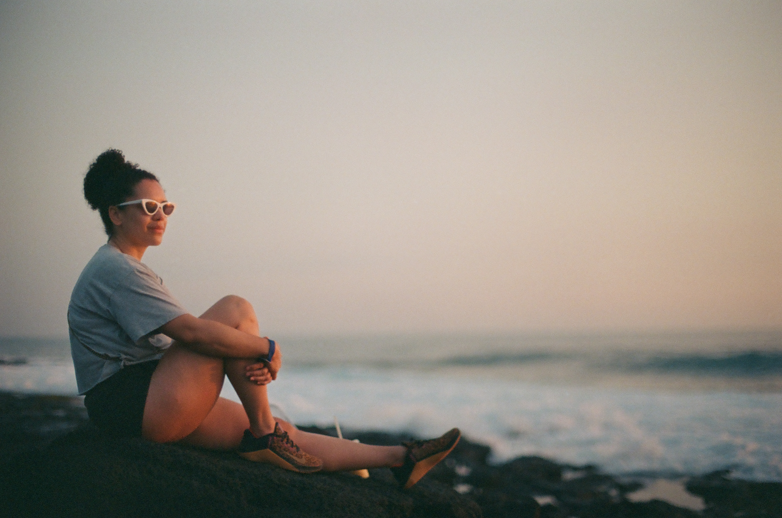 A person in white glasses and a gray shirt sits on dark rock with open ocean softly blurred behind.