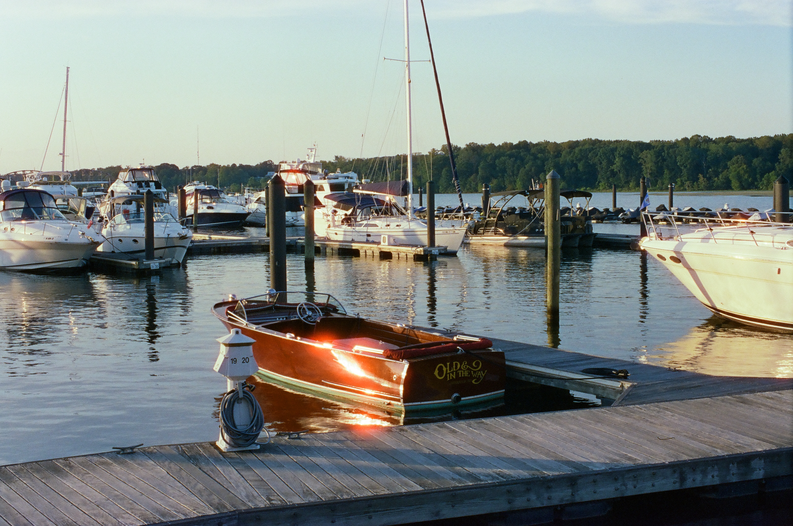 Wooden runabout lettered Old & In the Way tied near a post numbered 19 and 20, with moored powerboats, tall masts, and a green treeline under pale blue sky.