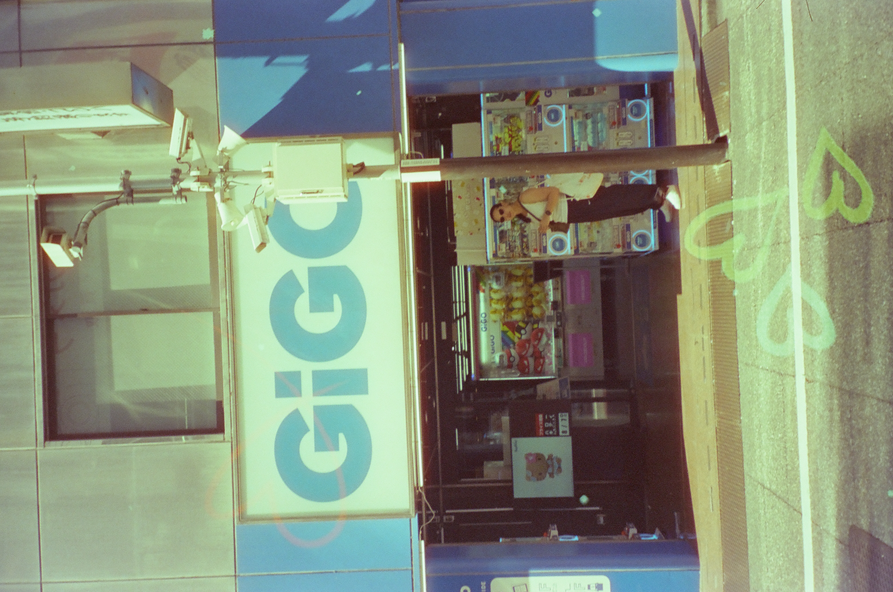 A person in sunglasses stands near a pole with security cameras outside a storefront with a large sign reading 'GiGO,' with green heart outlines on the crosswalk below.