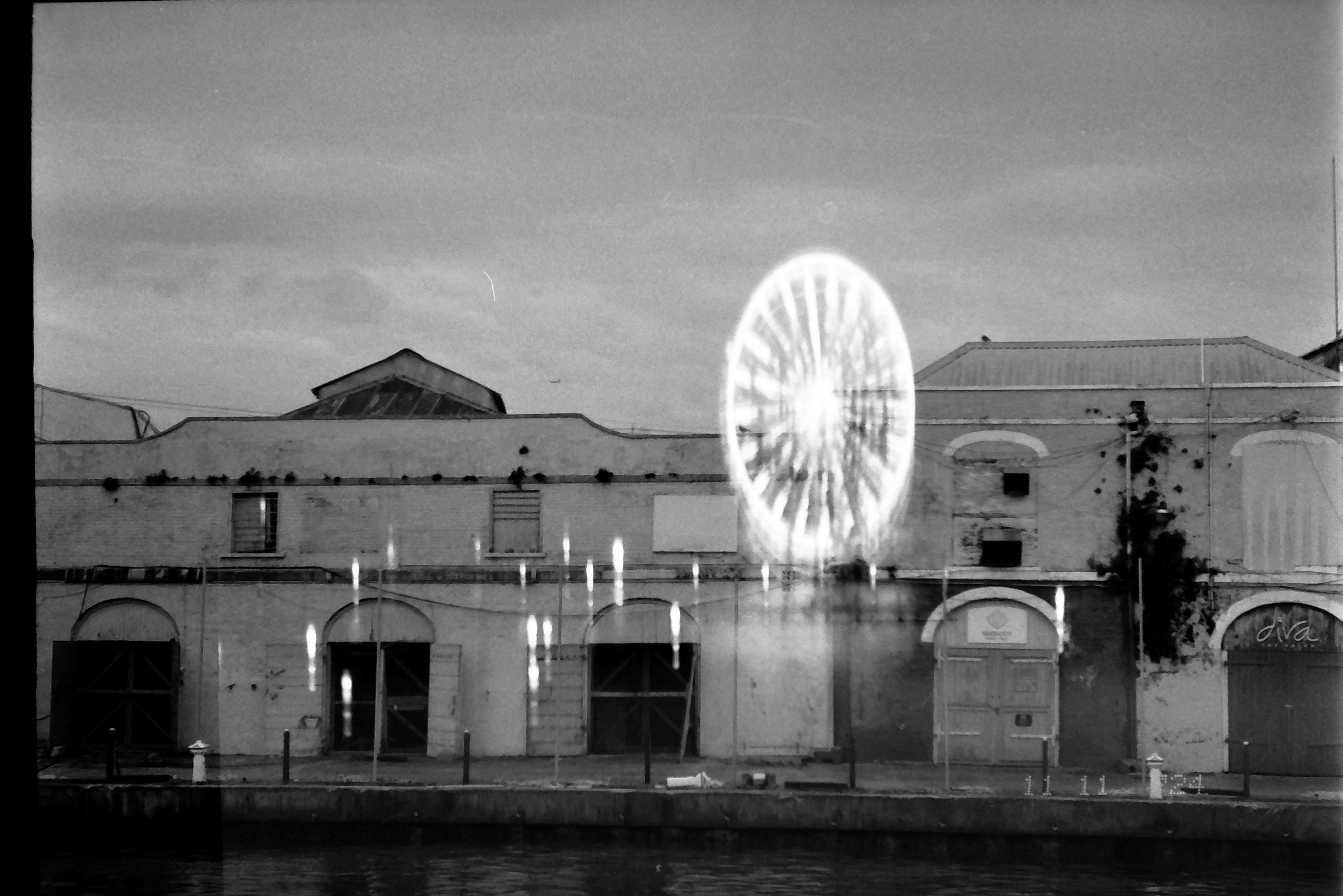 A blurred circular shape resembling a spinning wheel of light hovers above a row of low weathered buildings along a waterfront.