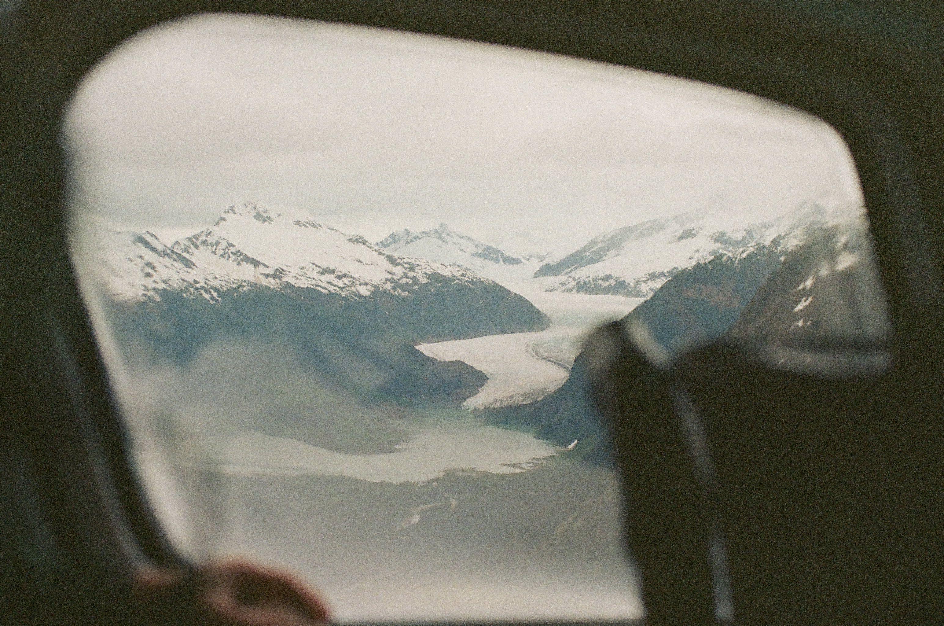 A rounded aircraft window frames snowy mountains, a long glacier, and pale water far below.