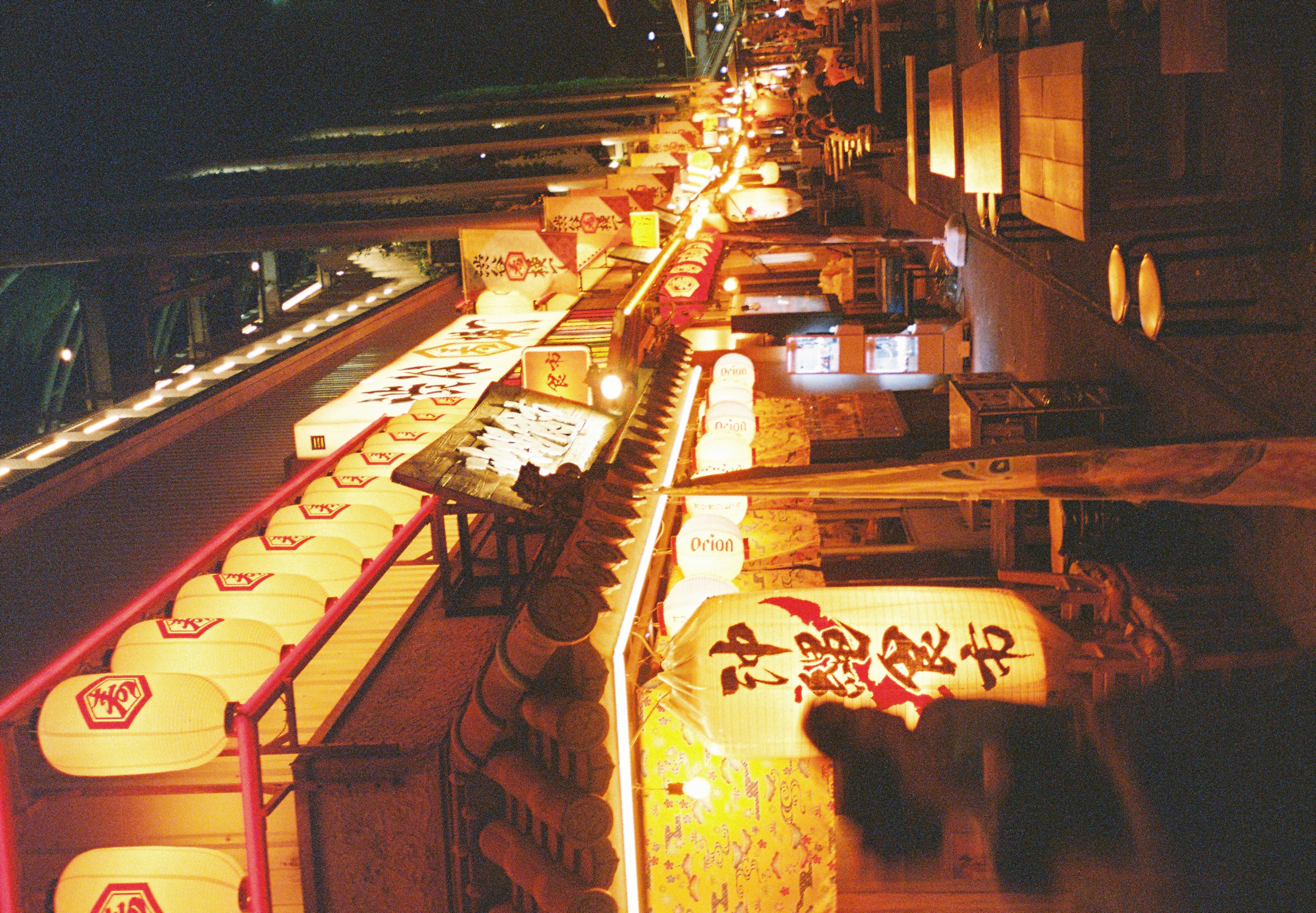 A covered alley at night lined with glowing white paper lanterns, red beams, and tiled eaves, with wooden tables and round stools receding along the right.
