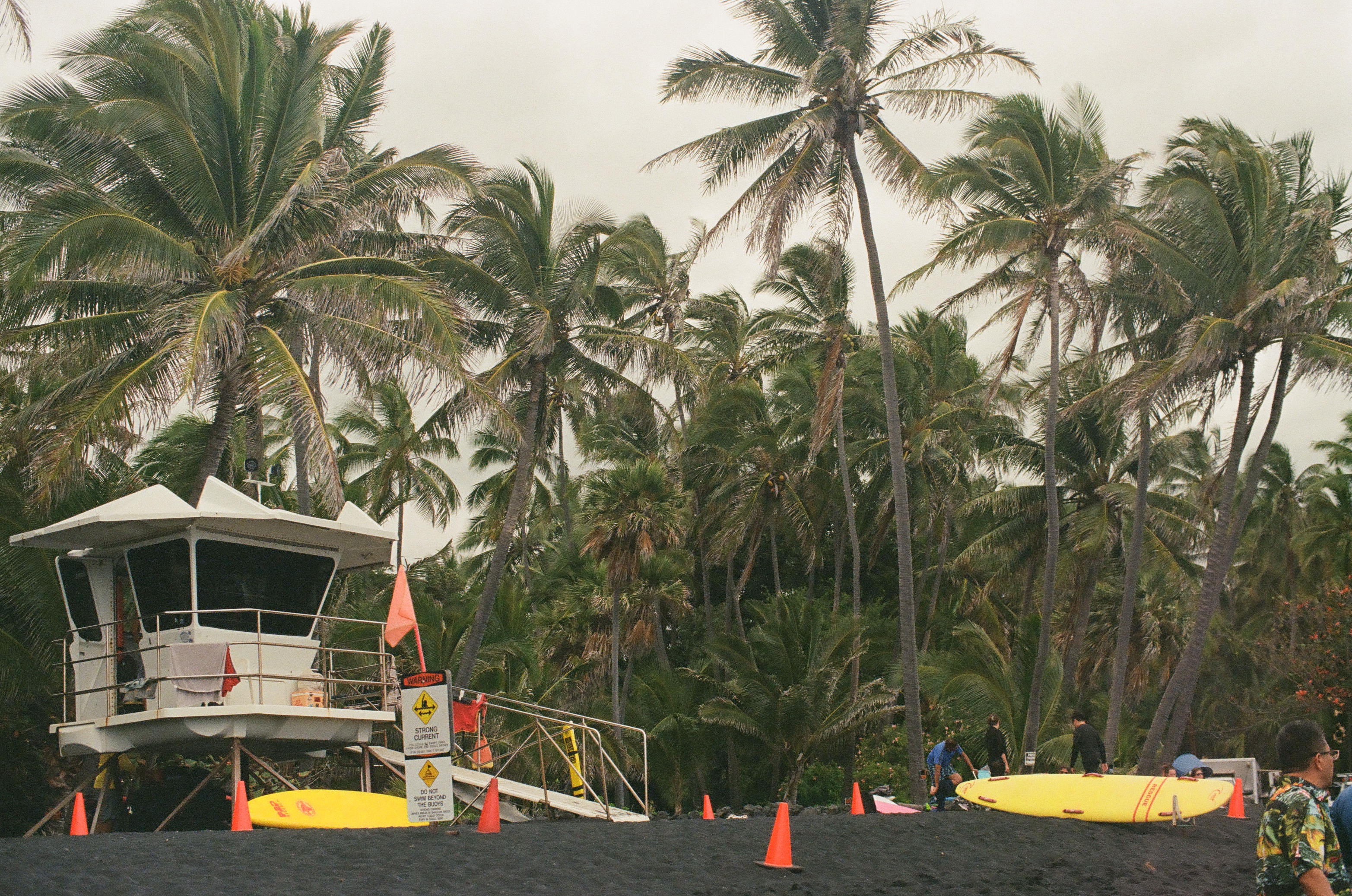 A white lifeguard tower with an orange flag stands beside a strong-current warning sign, orange cones, and yellow surfboards under tall palms.