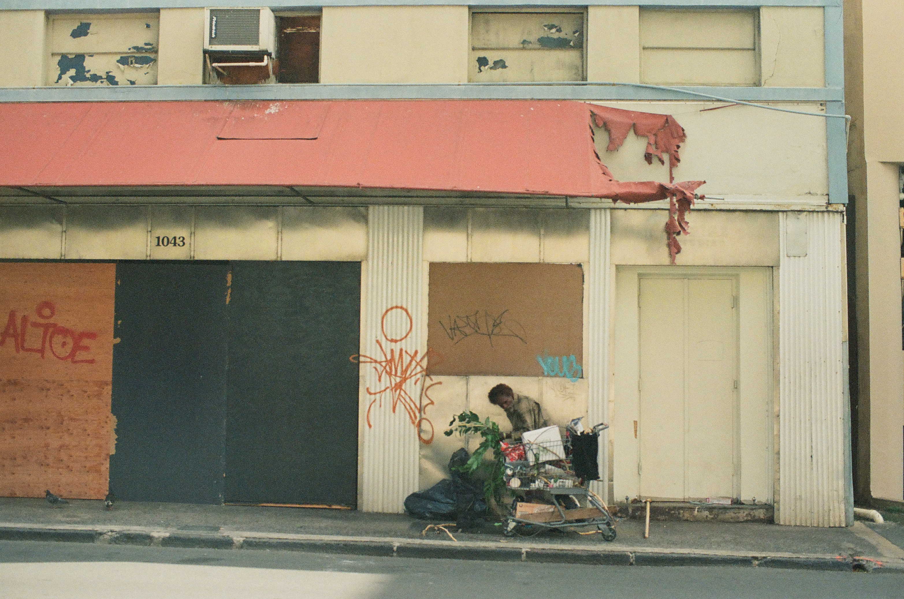 A person bends over a loaded shopping cart beside a boarded storefront with a torn red awning and graffiti.