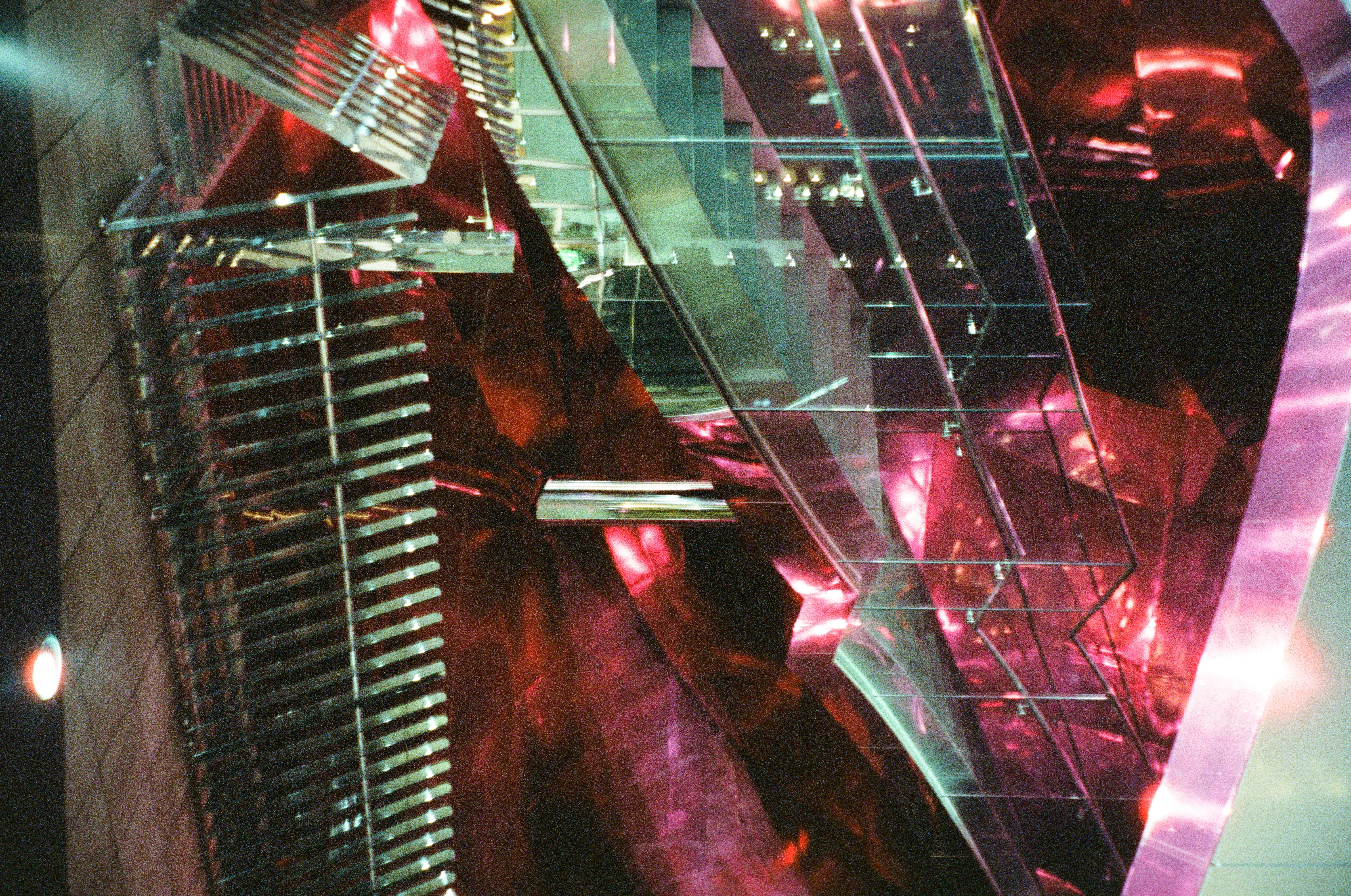 A staircase with glass railings descends past faceted, mirror-finished panels glowing magenta, with a chrome slatted screen at its foot.