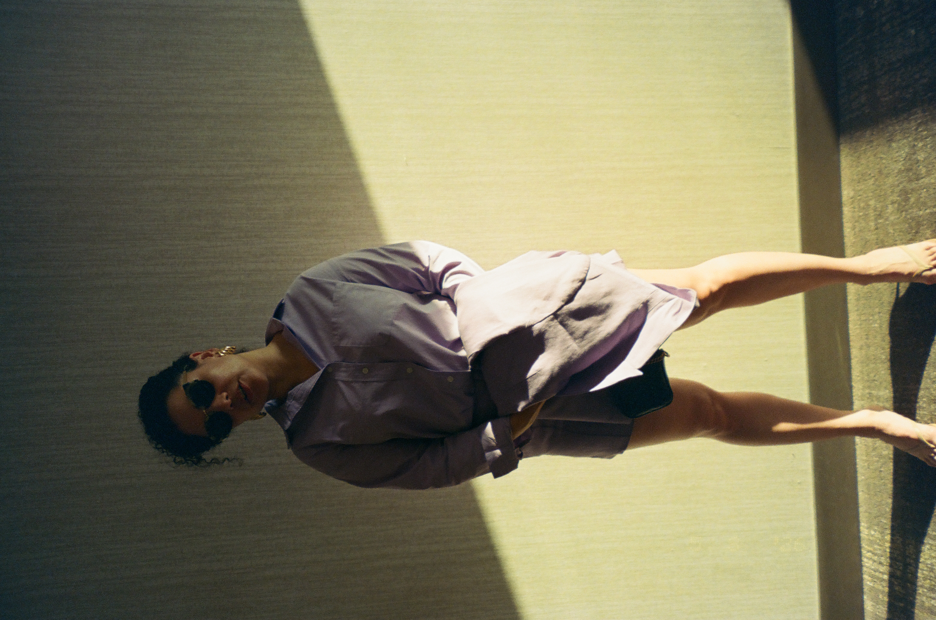 A person in dark sunglasses and a loose lavender-gray shirt stands against a wall divided diagonally into bright sunlight and shadow.