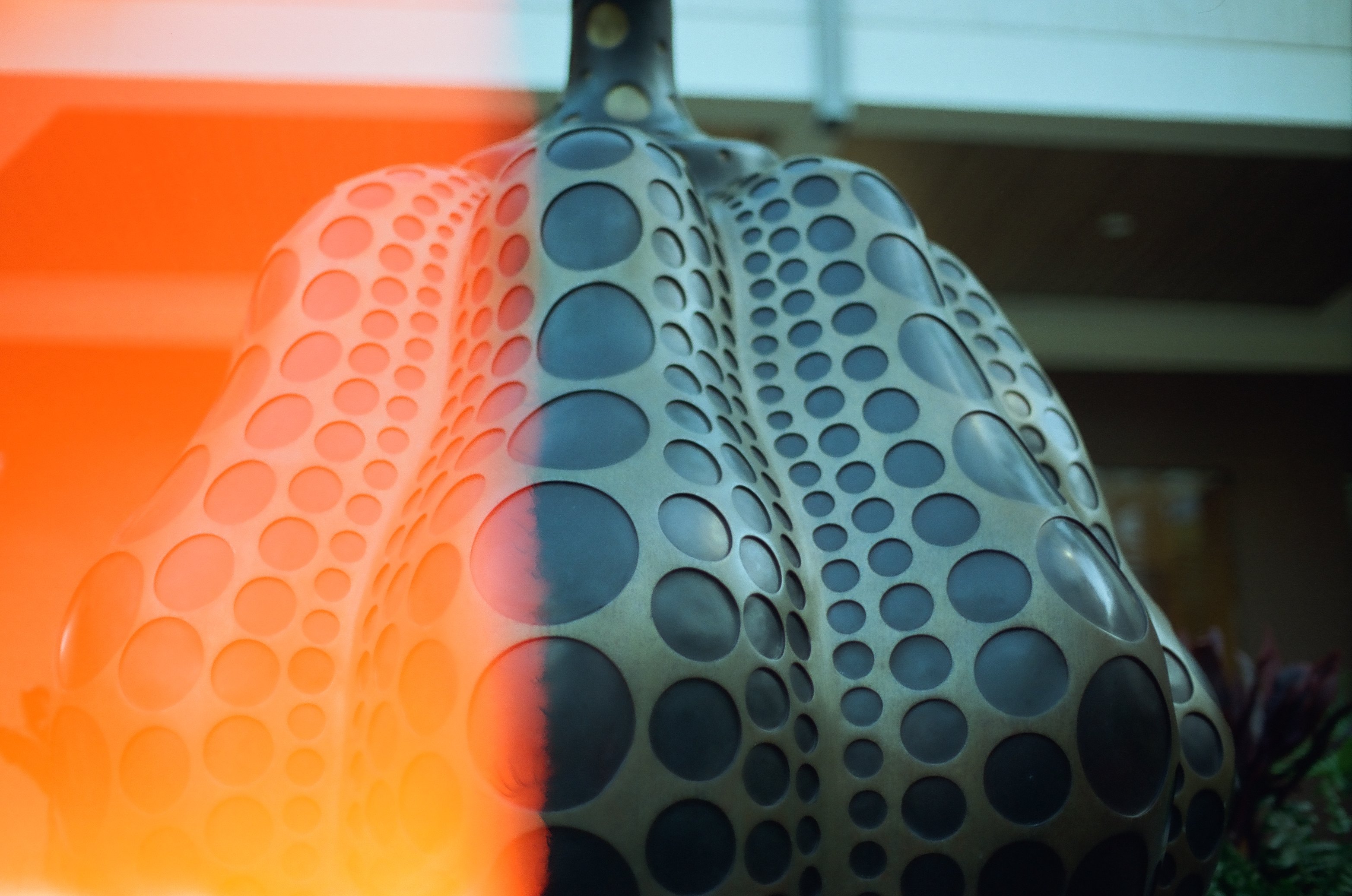A large gray perforated sculptural form fills the frame beneath a broad orange film light leak.