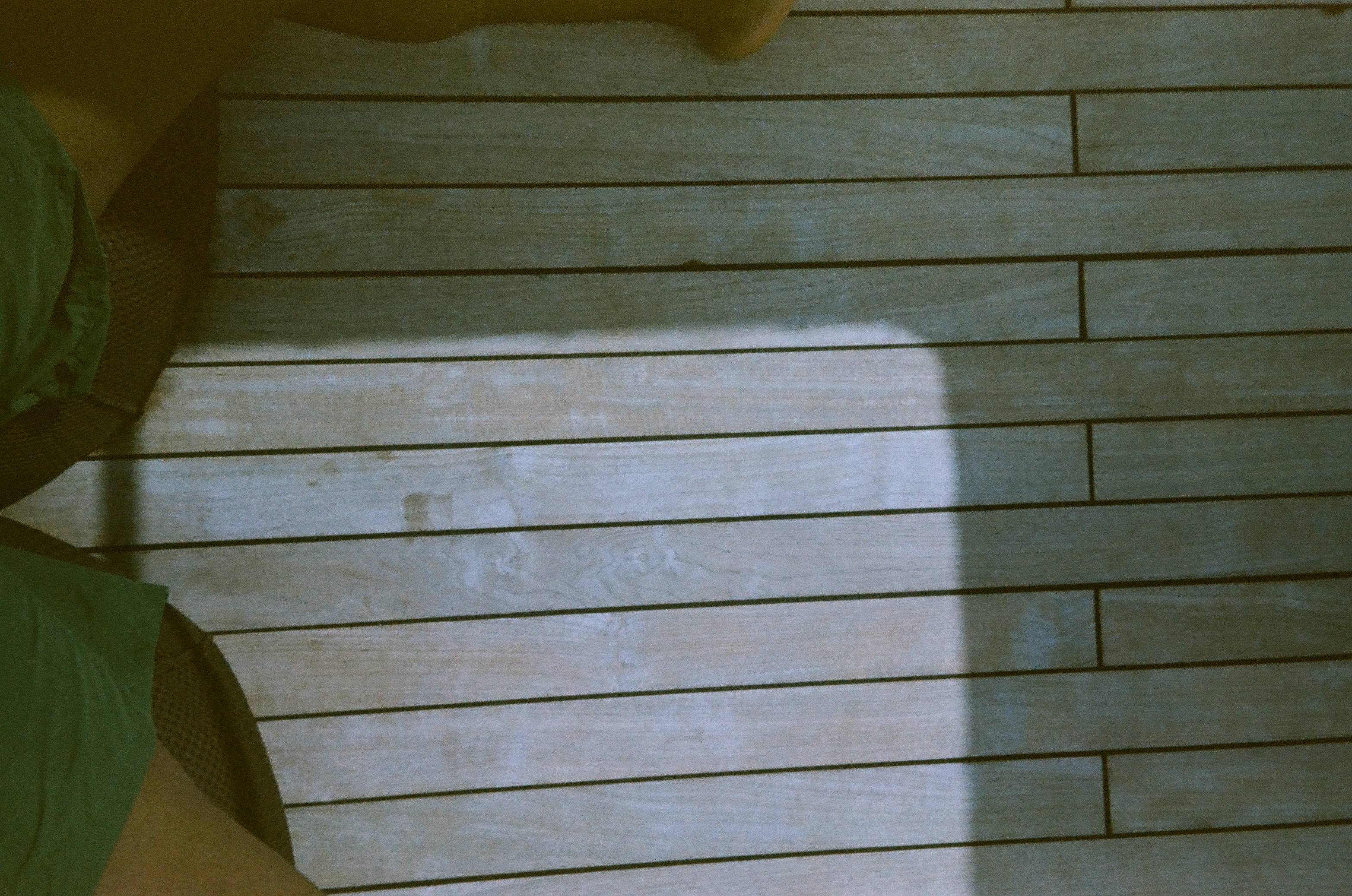 A broad rectangular shadow and cropped feet fall across pale wooden deck planks in strong sun.