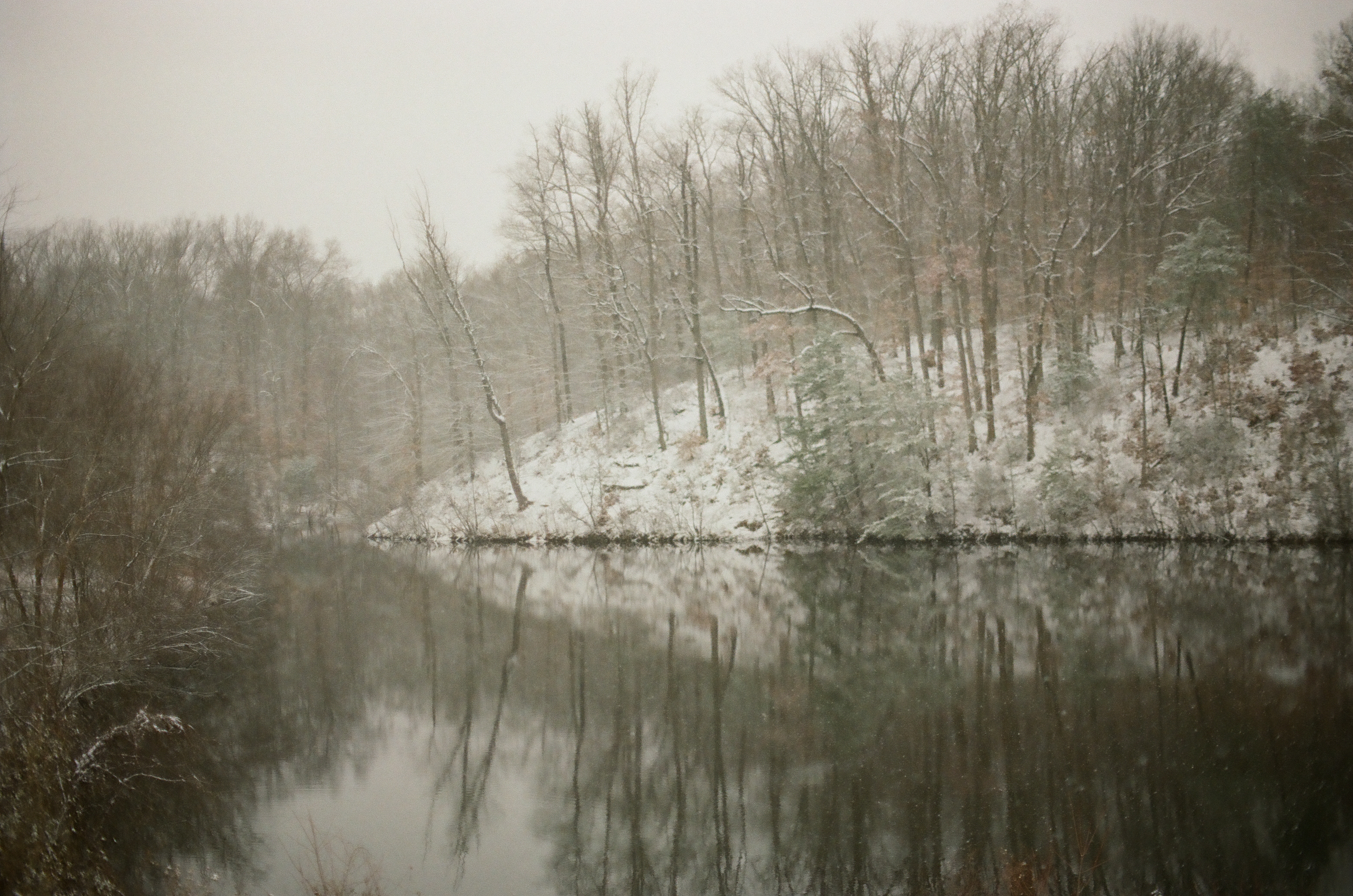 Bare trees and a snow-dusted wooded bank are mirrored in still dark water beneath a flat pale sky.