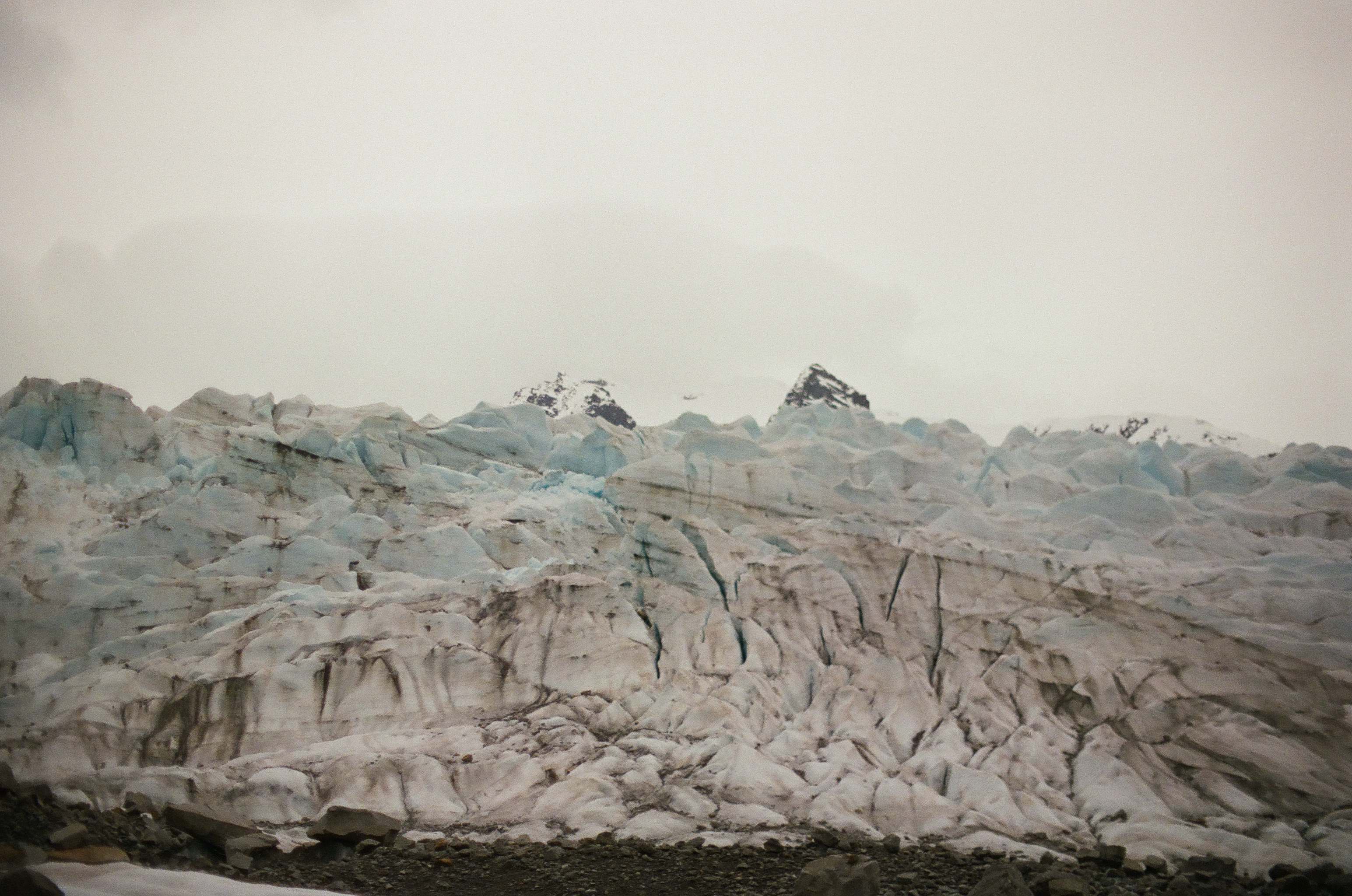 A broad glacier of ridges and crevasses reaches toward low snowy mountains beneath a blank sky.