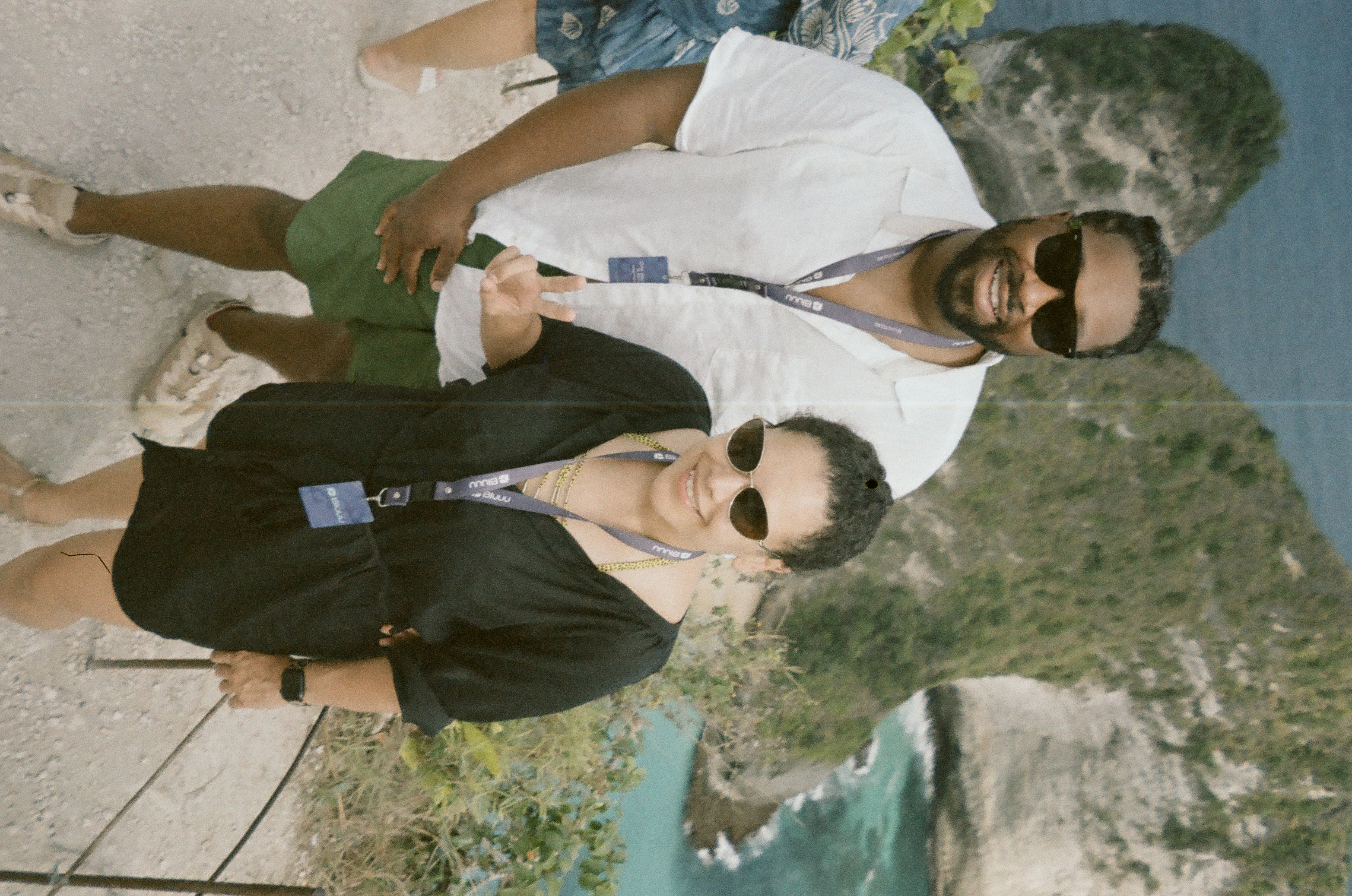 Two adults wearing sunglasses and lanyards face the camera above steep rock and blue-green water; one raises two fingers, and part of another person is cropped at left.